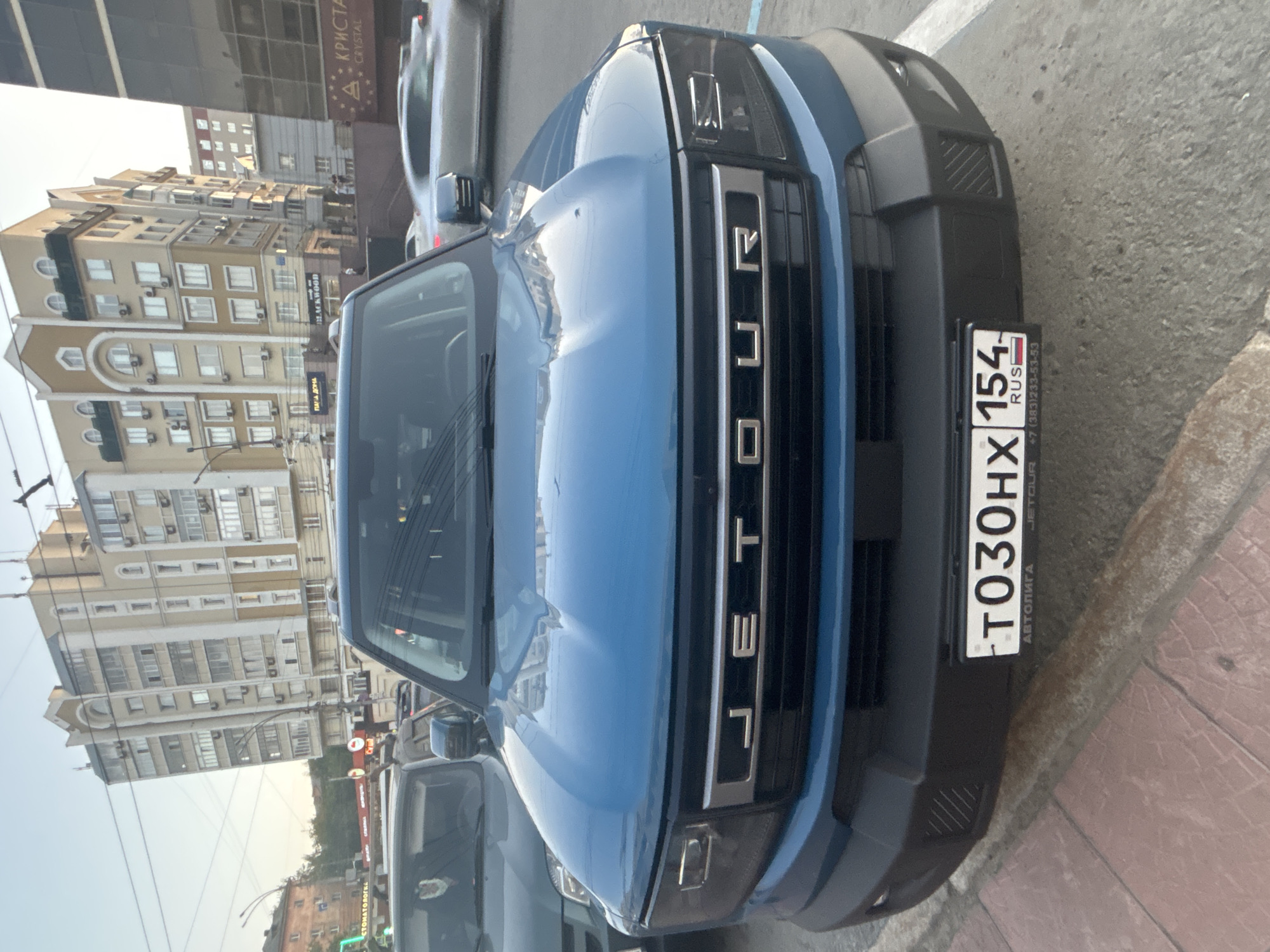 A blue JETOUR SUV is parked on a street in Novosibirsk, Russia. The license plate is visible and reads "T030HX 154". In the background, there are buildings, other cars, and utility lines. The sky is clear and the sun appears to be setting.