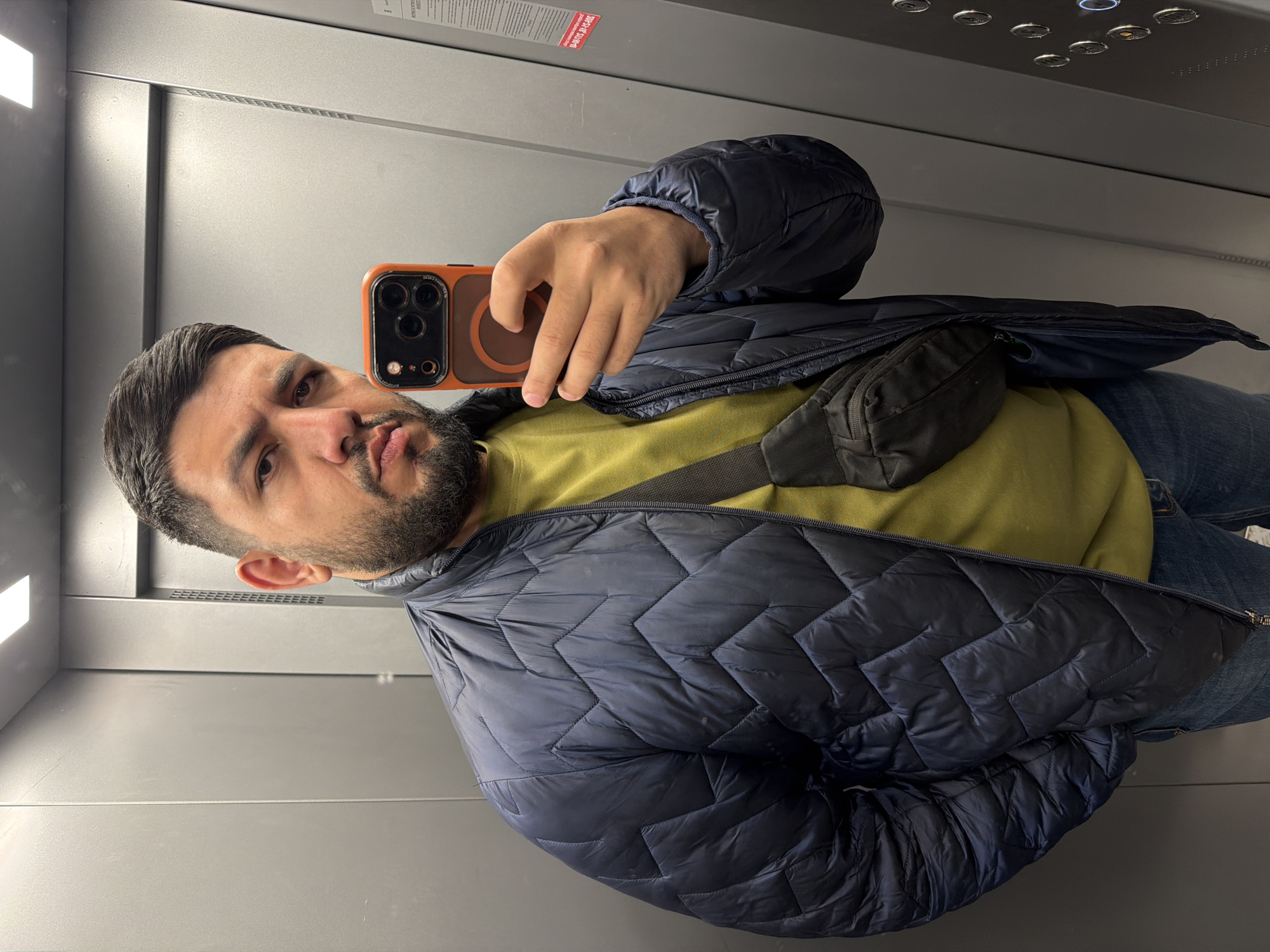 A man is taking a selfie in an elevator. He is wearing a blue puffer jacket over a green shirt, with blue jeans. He is holding an orange and black iPhone. The elevator has grey walls and metal control buttons at the top. There is no indication of the location being Krasnoyarsk, Russia.