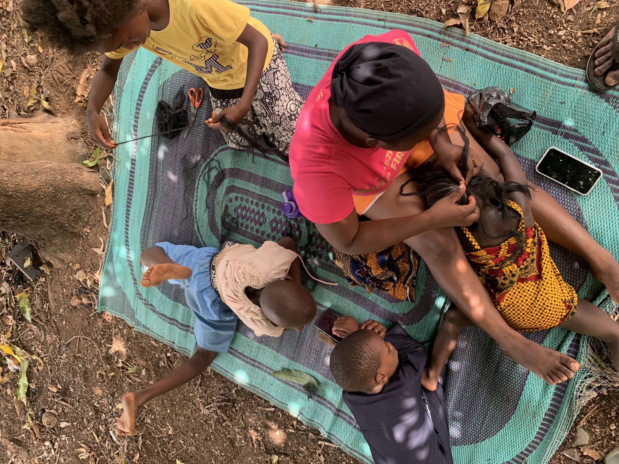 A high-angle shot depicts a group of people sitting on a patterned blue and green mat outdoors, possibly in a yard or park in Akwanga, Nigeria. An adult woman in a pink shirt and black head covering sits with her legs outstretched, braiding the hair of a child in a yellow and red patterned dress. A second child in a yellow shirt and printed pants sits nearby, manipulating black hair extensions with scissors. Two younger children are lying on the mat near the woman; one in a blue shirt, and another in a dark blue shirt, appearing to be interacting with a mobile phone. A second mobile phone, a black plastic bag, and what appears to be a small bottle are also on the mat. The surrounding ground is dirt and loose leaves, and a tree trunk is visible in the upper left corner of the shot.