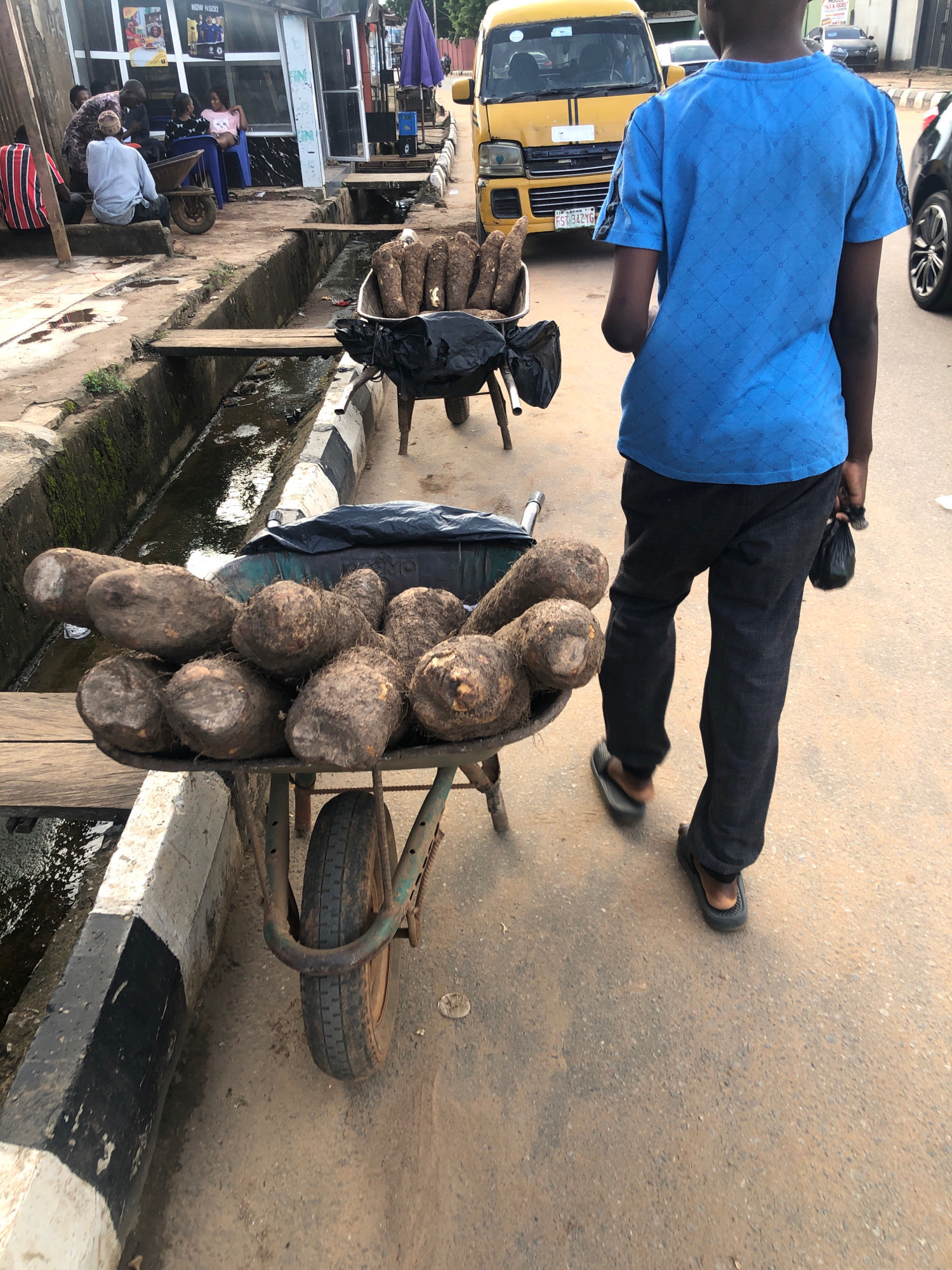 The image depicts a street scene in Lagos, Nigeria, with a young person wearing a blue t-shirt and black pants walking on a paved road. In the foreground, a wheelbarrow filled with yams is positioned. The wheelbarrow's contents are partially covered by a black tarp. To the left of the wheelbarrow is a drainage ditch with murky water and mossy concrete walls. A narrow wooden plank bridges the ditch. In the background, a yellow van is parked, and other pedestrians and structures are visible, suggesting a commercial or residential area. The overall environment appears to be an outdoor marketplace or roadside.