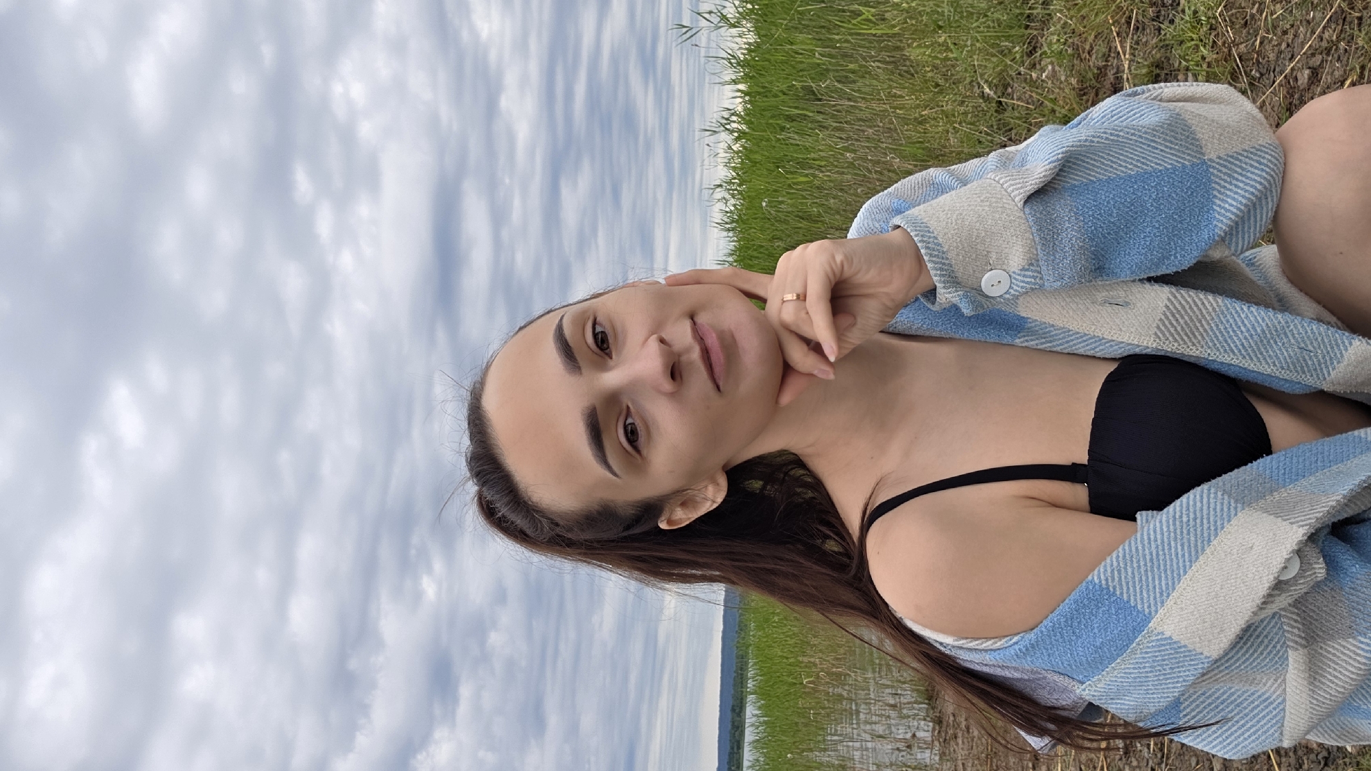 A young woman with dark brown hair pulled into a ponytail is captured in a medium shot, posing outdoors. She is wearing a black bikini top and a light blue and white checkered open shirt. Her right hand rests against her chin, and she wears a gold ring on her ring finger. The background consists of tall green reeds bordering a body of water, with a distant shoreline visible under a cloudy sky. The scene appears to be set in a natural environment, possibly near a lake or river.