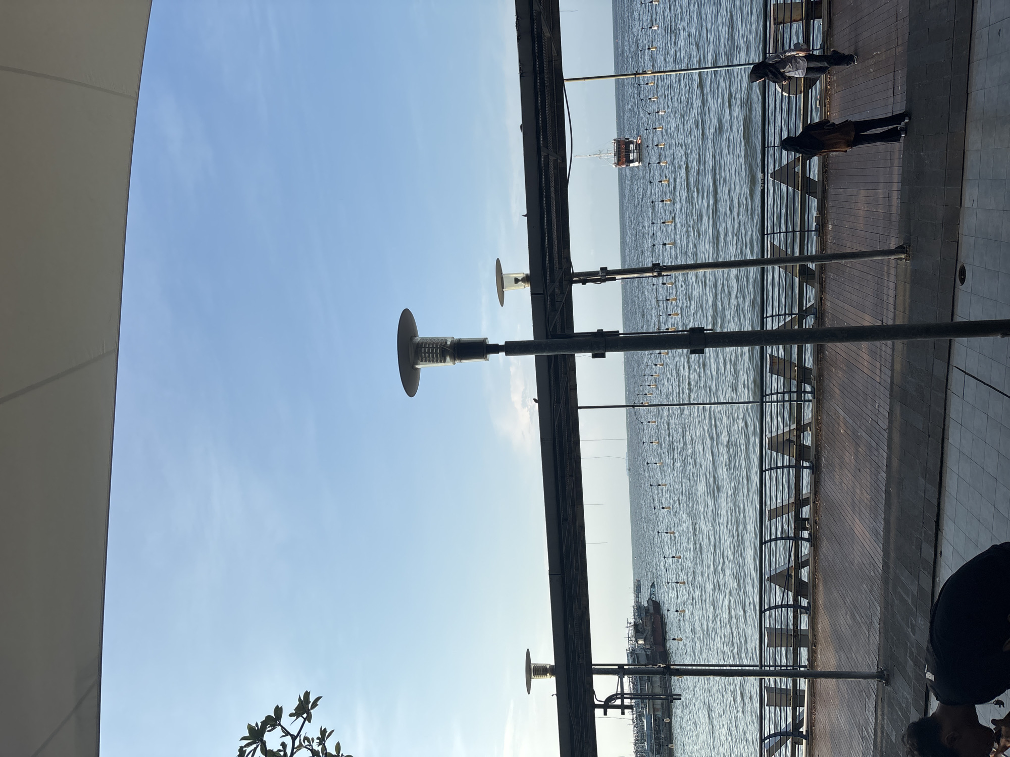The image shows an outdoor scene in North Jakarta, Indonesia, depicting a waterfront view. The sky is partly clear. The body of water is visible behind a metal structure and several evenly spaced poles, possibly light poles. Two people can be seen in the upper right of the frame, standing on a wooden walkway. Additionally, another person is visible near the bottom right corner. A white awning is visible on the left side of the frame, and some foliage is visible at the bottom left corner. In the water, a platform or structure is located in the background, appearing near a line of vertically suspended lights or buoys. Several boats are also visible.