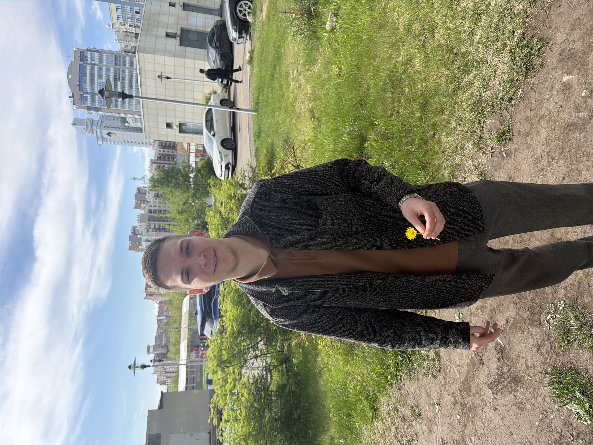 A young man stands outdoors in Krasnoyarsk, Russia, holding a dandelion in his right hand and a cigarette in his left. He is wearing a brown shirt with a dark gray cardigan and gray pants. In the background, there are buildings, trees, cars, streetlights, and a cloudy sky. The ground is a mix of grass and dirt. A street is also visible with cars and what appears to be a person crossing it.