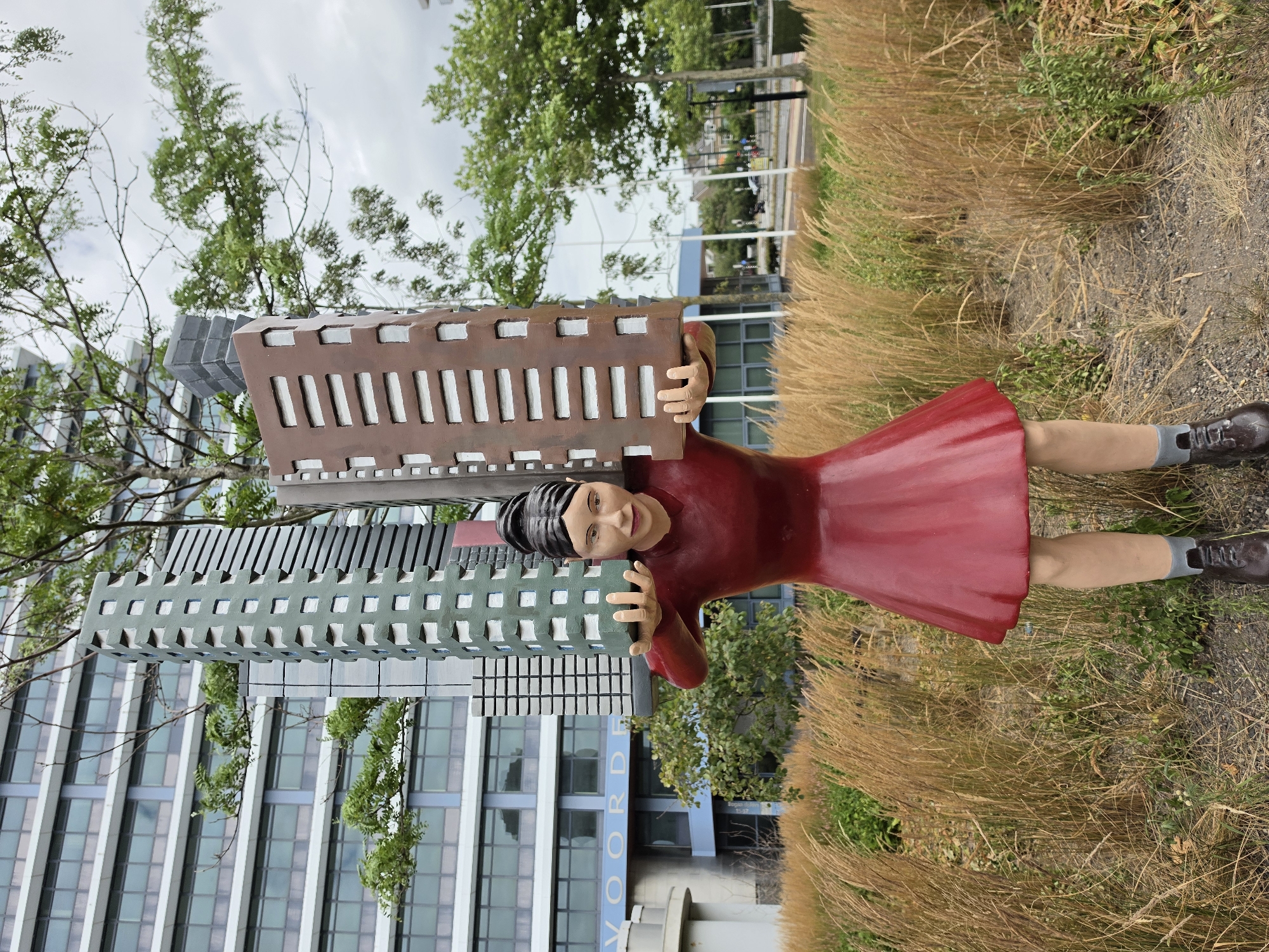 This image shows a statue of a young woman wearing a red dress and dark shoes, carrying models of several skyscrapers on her shoulders. She is standing in a grassy area with tall, dry reeds in the foreground and a modern building in the background. The building, which appears to be an office building, has a facade of light-colored panels and blue-tinted windows. The statue is positioned in front of the building, which is located at the intersection of Herenstraat and Van Duivenvoordestraat in Rijswijk, Netherlands. Trees with green leaves are visible behind the statue and in front of the building. The sky is overcast.
