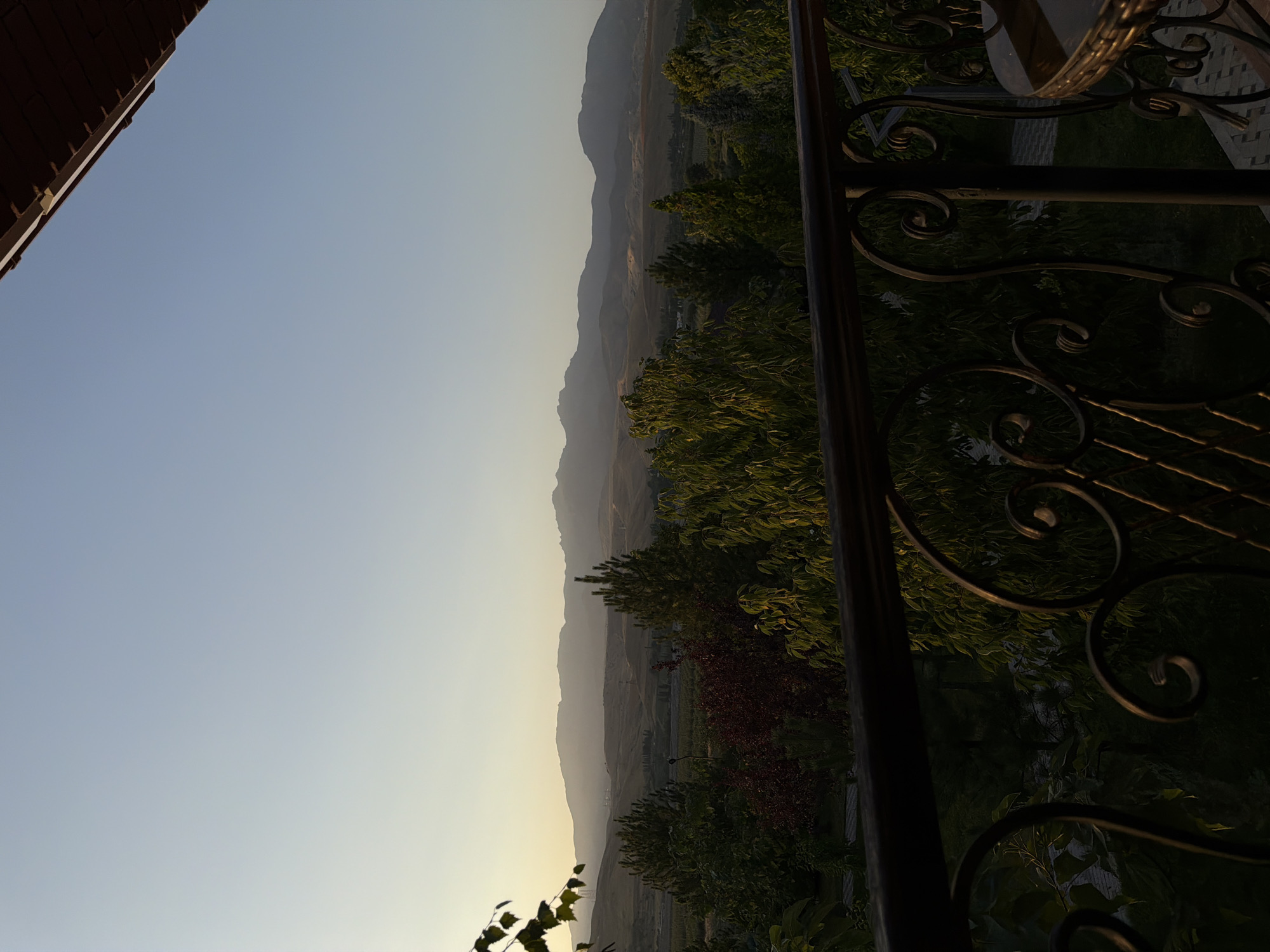 The image shows a view of a landscape from a balcony in Karankultugay, Uzbekistan. The foreground features a dark-colored metal railing with decorative scrollwork. Beyond the railing, a lush green and red-leafed garden is visible, with various trees and foliage. In the middle ground, there appears to be a collection of buildings and agricultural fields, possibly vineyards, stretching towards the horizon. The background is dominated by rolling hills and distant mountains, silhouetted against a pale, hazy sky that suggests either early morning or late evening light. The overall impression is one of a serene, rural or semi-rural setting.