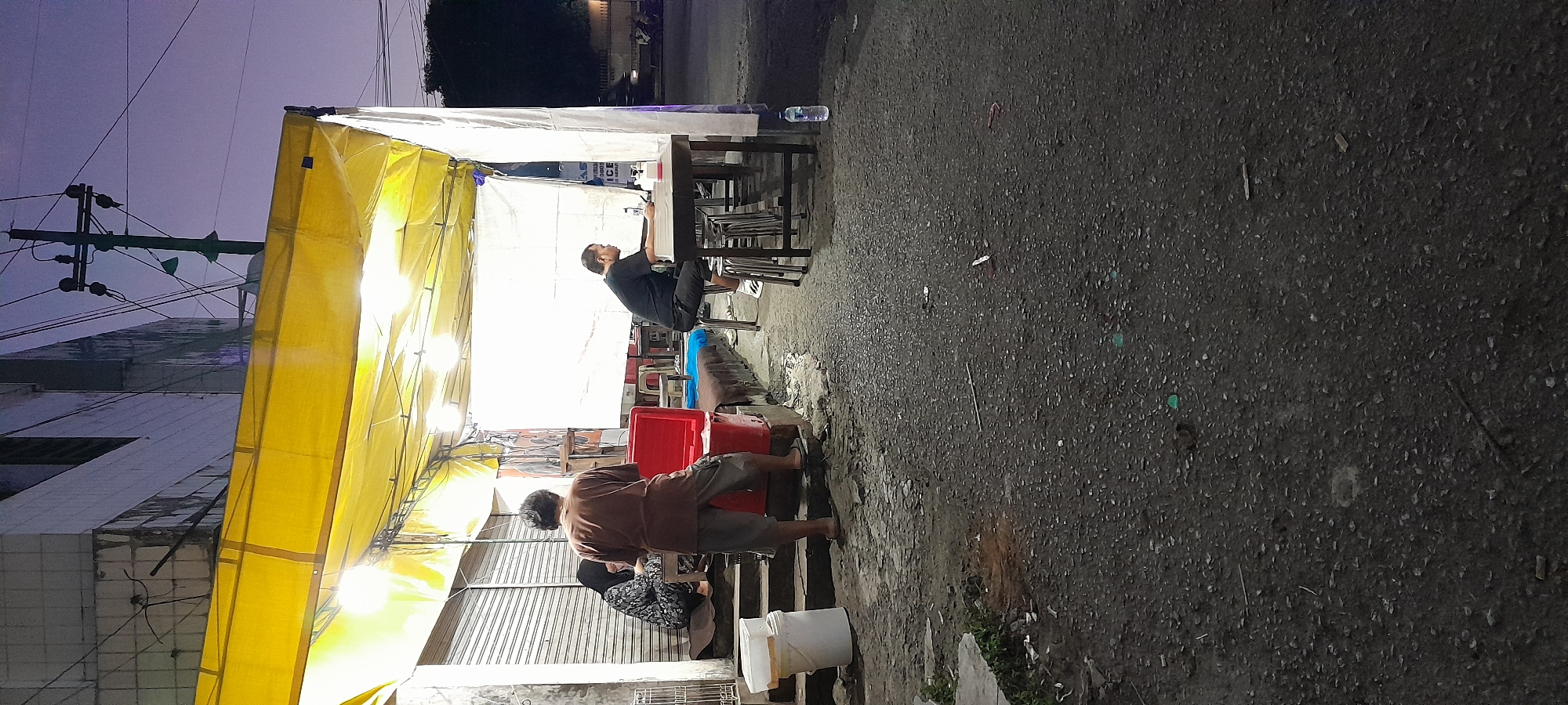 In Rogojampi, Indonesia, an outdoor food stall is set up under a yellow awning. The stall is illuminated by string lights. In the foreground, a man wearing a brown t-shirt and grey shorts stands facing away from the camera, looking towards the stall. He is positioned on a step. To his left, a woman in a patterned headscarf and dress sits at a table, looking down. A red cooler is placed in front of the steps. To the right, another man in a black t-shirt sits at a table with several stools, looking at his phone. The ground is paved and appears somewhat rough, with scattered debris. A plastic bottle of water rests on the ground near the table. The scene takes place during twilight.