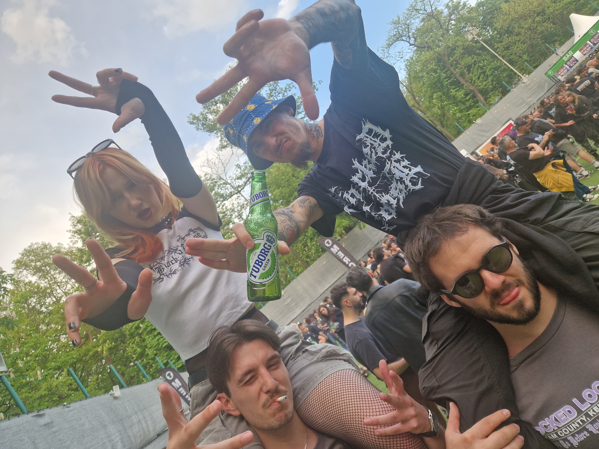 This image, taken at an outdoor event, possibly a music festival, shows four individuals posing for a photograph. The foreground features a young woman with orange-tinted hair, wearing a white t-shirt, black arm warmers, and sunglasses perched atop her head. She is extending both hands towards the camera, fingers spread in a gesture. Adjacent to her is a man wearing a black t-shirt, blue bucket hat, and multiple tattoos on his arms. He holds a green Tuborg beer bottle and also makes a hand gesture towards the camera. Below them, a man with dark hair and a mustache sits on someone's shoulders, a cigarette in his mouth, and his fingers are posed in a similar gesture. A fourth man, wearing sunglasses and a dark t-shirt, sits on the shoulders of a person behind him, appearing to be a similar situation to the others. The background is filled with a dense crowd of people, tents, trees, and signs, suggesting a festival or concert atmosphere. The sky is partially visible with clouds.