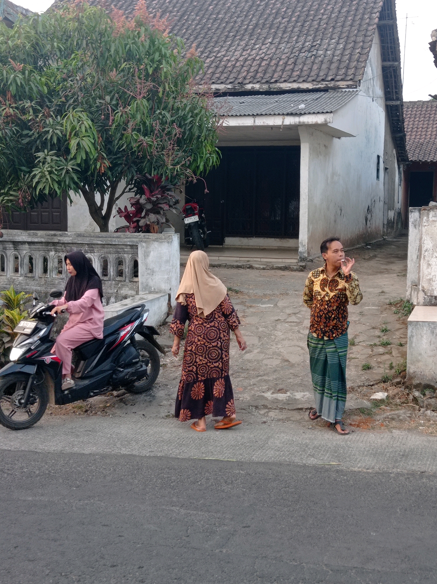 The image depicts an outdoor scene in front of a building, likely a house, in Purwosekar, Indonesia. A woman wearing a pink headscarf and matching pink attire is seated on a black scooter. To her right, another woman in a brown headscarf and a patterned brown and orange dress stands facing away from the camera. A man in a batik shirt and a green and white striped sarong stands to the right of the second woman, holding a cigarette in his right hand and appearing to smoke it. The scooter is parked on a paved surface, and behind the people, a wall with decorative elements separates the area from the house. A tree with green leaves and reddish flowers is visible to the left of the frame, and the building features a tiled roof and a white facade. The ground is a mix of paved areas and dirt.