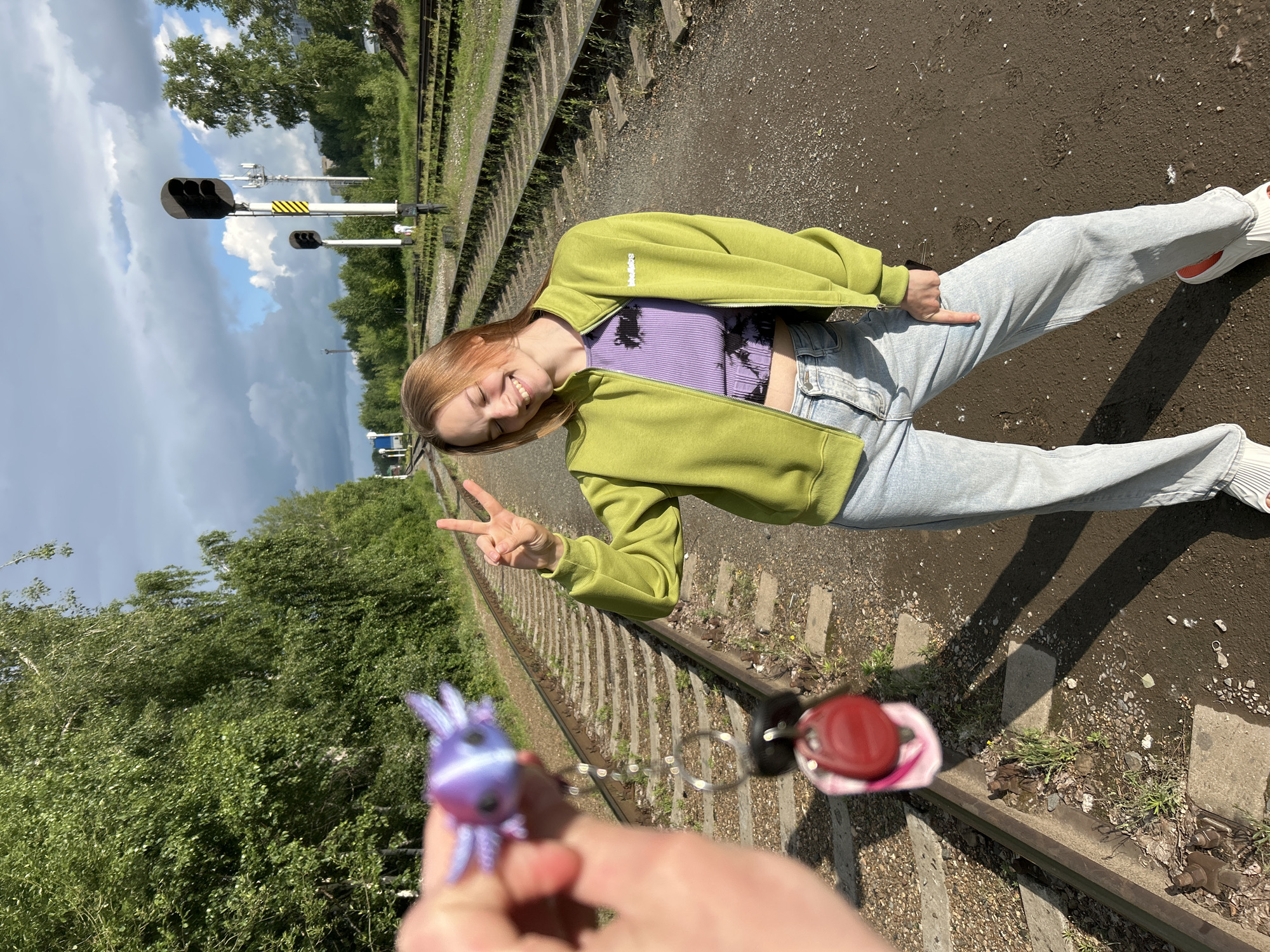 A young woman with blonde hair stands on a railway track in Chaikovsky, Russia, smiling and holding up two fingers in a peace sign. She is wearing a light green zip-up jacket over a purple crop top and light blue jeans. In the foreground, a hand holds up a keychain with a purple axolotl charm and red fob. The railway tracks stretch into the distance, flanked by trees and railway signals. The sky is partly cloudy.