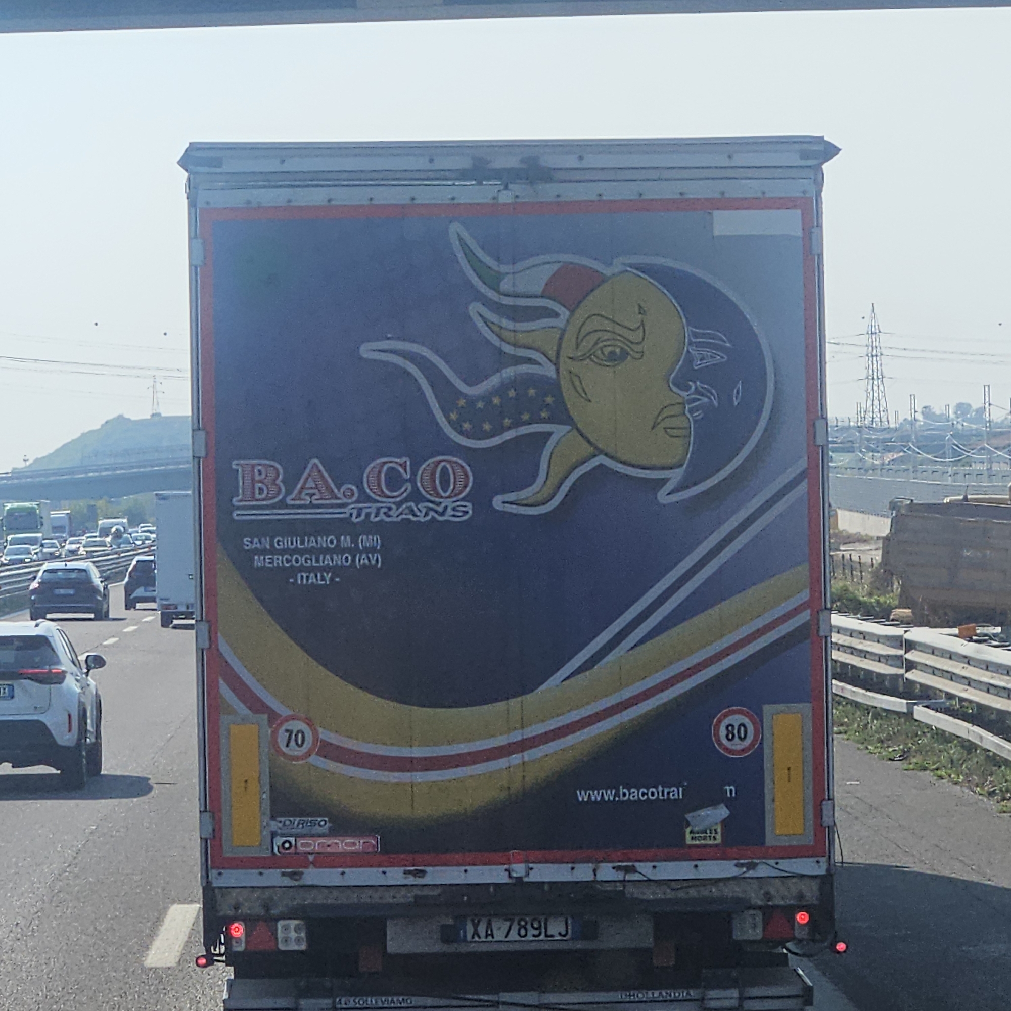 The image shows the rear of a large truck with a blue trailer, featuring a prominent graphic of a sun and moon with faces. Text on the trailer reads "BA.CO TRANS SAN GIULIANO M. (MI) MERCOGLIANO (AV) - ITALY -". The truck is driving on a highway with other vehicles visible in the background, indicating a traffic scene. There are speed limit signs of "70" and "80" on the trailer. The license plate of the truck is "XA 789LJ". The environment appears to be a sunny day on an Italian highway, possibly near Lonato del Garda, Italy, suggested by the overall context of Italian road signs and truck markings.