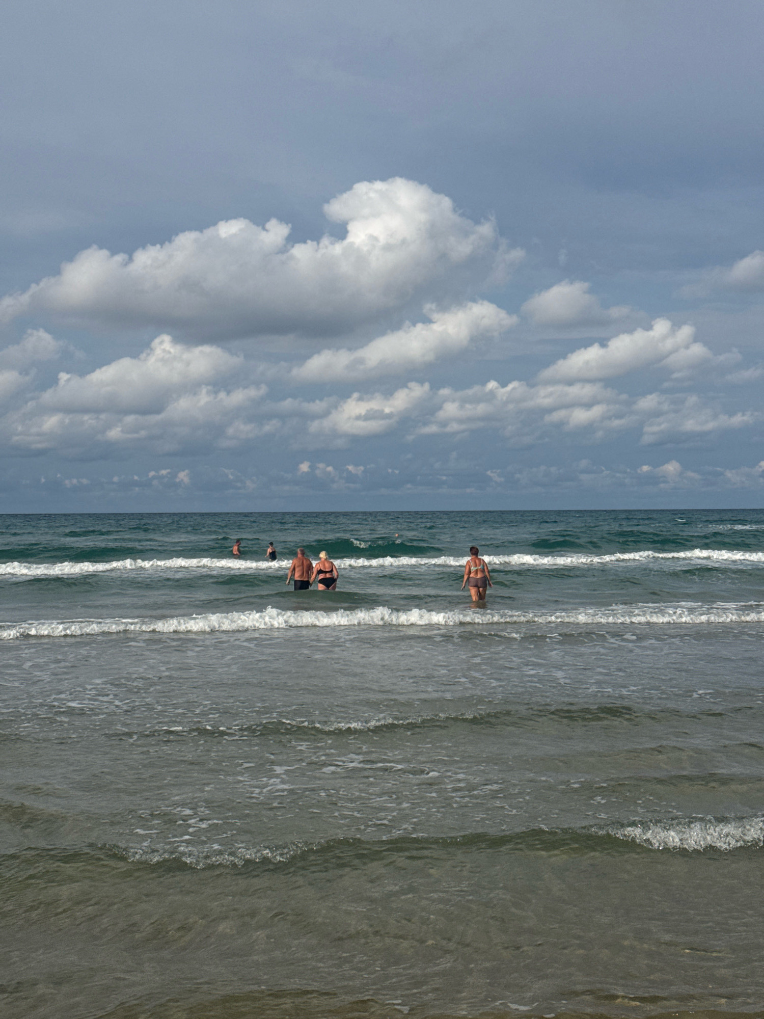 In Anapa, Russia, on a cloudy day, a group of people are wading in the sea. The water is a deep green, with visible waves breaking and receding towards the sandy shore. In the foreground, a couple stands in the water, with the man wearing dark swim trunks and the woman in a black bikini. Further out, a woman in a brown bikini is alone in the water. In the distance, two more individuals are visible, also wading in the sea. The sky is filled with dramatic, grey clouds, hinting at possible rain.