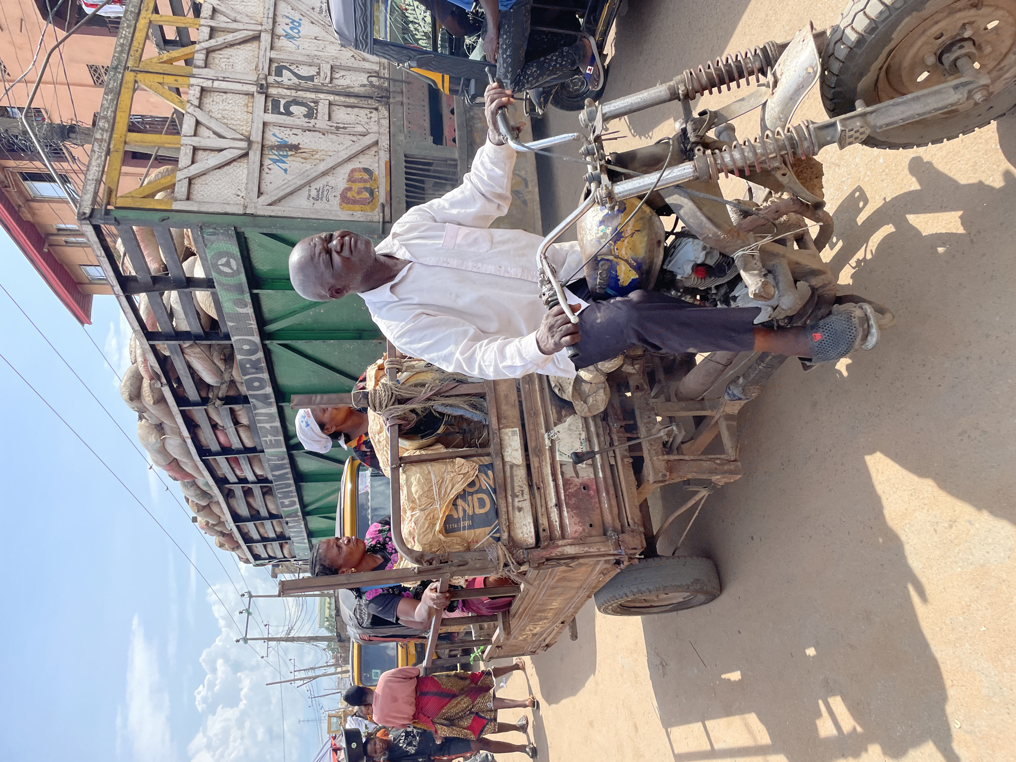 This image captures a bustling street scene under a bright, clear sky in the city of Unknown, Unknown. In the foreground, an elderly bald man, wearing a white long-sleeved shirt and dark pants, operates a weathered, custom-built motorized tricycle. His hands grip the handlebars, and he looks intently forward. The tricycle consists of a motorcycle front section fused with a rusty, metal cargo bed at the rear.
Seated within the tricycle's cargo bed are two women. The woman on the left wears a dark patterned top and a red skirt, looking towards the left side of the frame. Beside her, the second woman wears a white headscarf and a patterned top, also looking left. Two large, brown sacks, possibly containing goods, are visible next to them in the cargo bed. The tricycle itself shows significant wear, with visible rust, exposed engine components, and a faded multi-colored fuel tank. Its front wheel has a knobby tire, and the suspension springs are visible.
Behind the tricycle, a large, green cargo truck, heavily laden with numerous beige sacks, moves or is parked. The truck's wooden or metal side rails are filled to capacity, and partial lettering "AMARA CHUKWU EZU..." and "Nov 57 Mod. GD" are visible on its body. Further into the background, several yellow motorized tricycles (auto-rickshaws) are present, along with various pedestrians, including a woman balancing a dark object on her head and another in a pink top walking away.
The road surface is unpaved, appearing as light brown dirt or sand, with distinct shadows indicating strong overhead sunlight. In the background, multi-story buildings with faded paint, including an orange-hued structure with a red roofline, line the street. Numerous utility poles with a dense tangle of overhead wires are visible against the blue sky, which features scattered white clouds. The overall scene depicts active transportation and commerce in a busy, urban environment.
