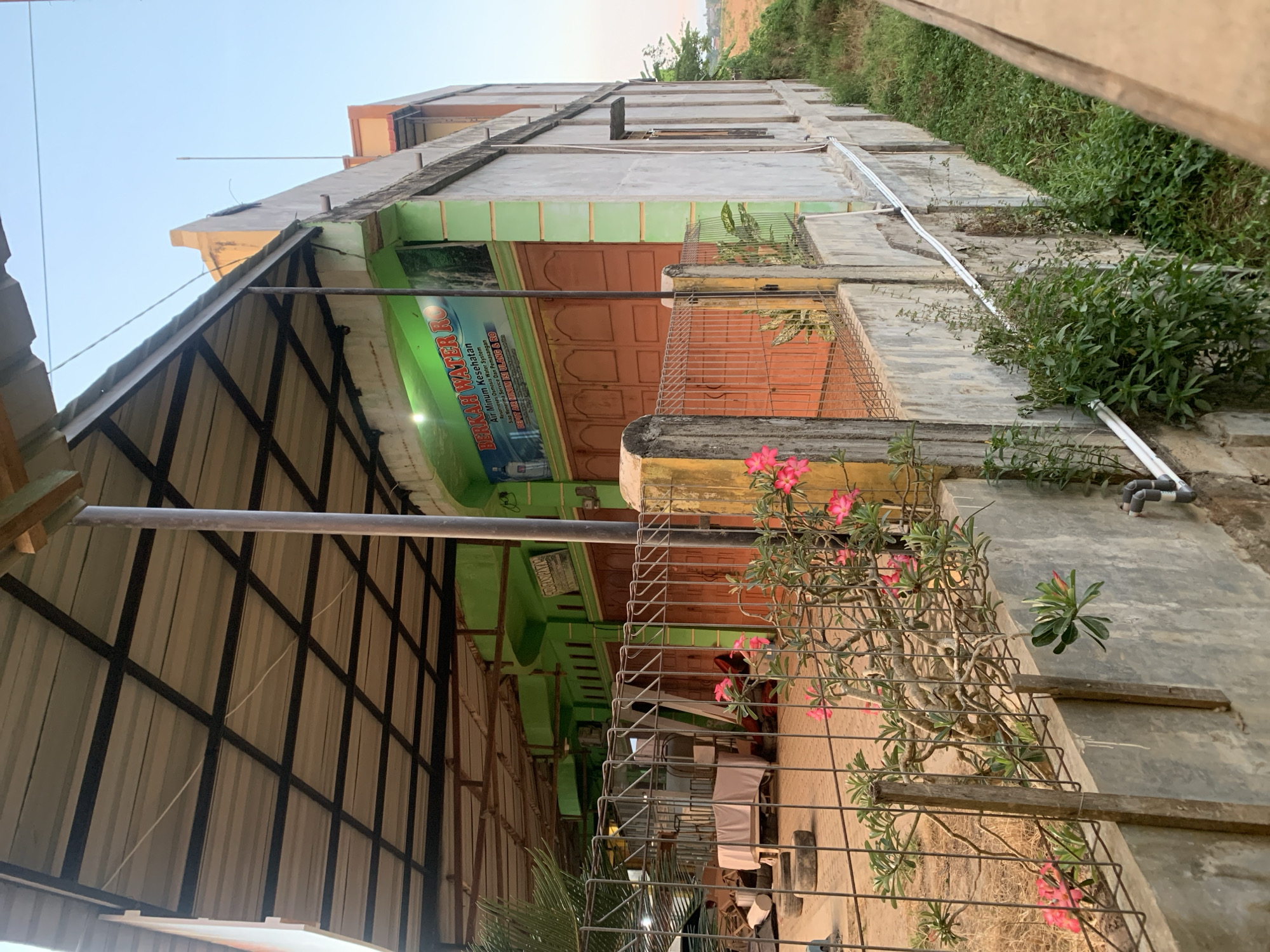 The image shows a roadside building with a green facade and orange doors, likely a commercial establishment. A sign on the building reads "BERKAH WATER RO Air Minum Kesehatan Osmosis Water System". Pink flowers are visible in the foreground, growing in front of a metal fence. The building appears to be in a rural or semi-urban area, possibly in Kualadua, Indonesia, given the presence of lush vegetation and a clear sky. The overall scene suggests a business operating in a warm climate.