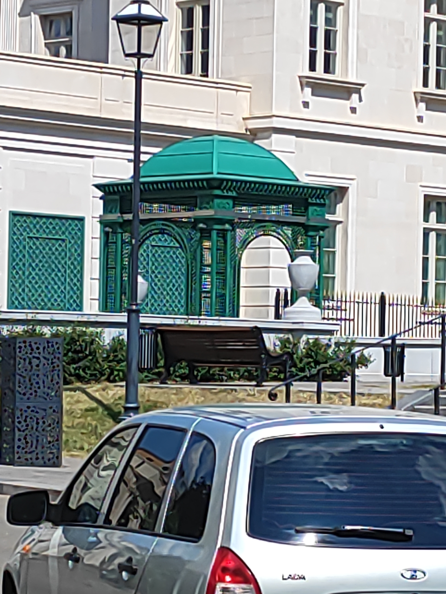 The image depicts a scene in Moscow, Russia, featuring a building with classical architecture, characterized by its white stone facade and large windows. In front of the building stands a decorative green pavilion with intricate latticework and a domed roof. A lamppost with a black pole and a white lantern is positioned to the left of the pavilion. To the right of the pavilion, a white urn-like sculpture rests on a pedestal. A wooden bench with black metal legs is placed in front of the pavilion, surrounded by greenery. A black metal fence runs along the right side of the scene. In the foreground, the rear portion of a silver car, identified by the "LADA" logo on its trunk, is visible. The car's rear window has a wiper and horizontal blinds.