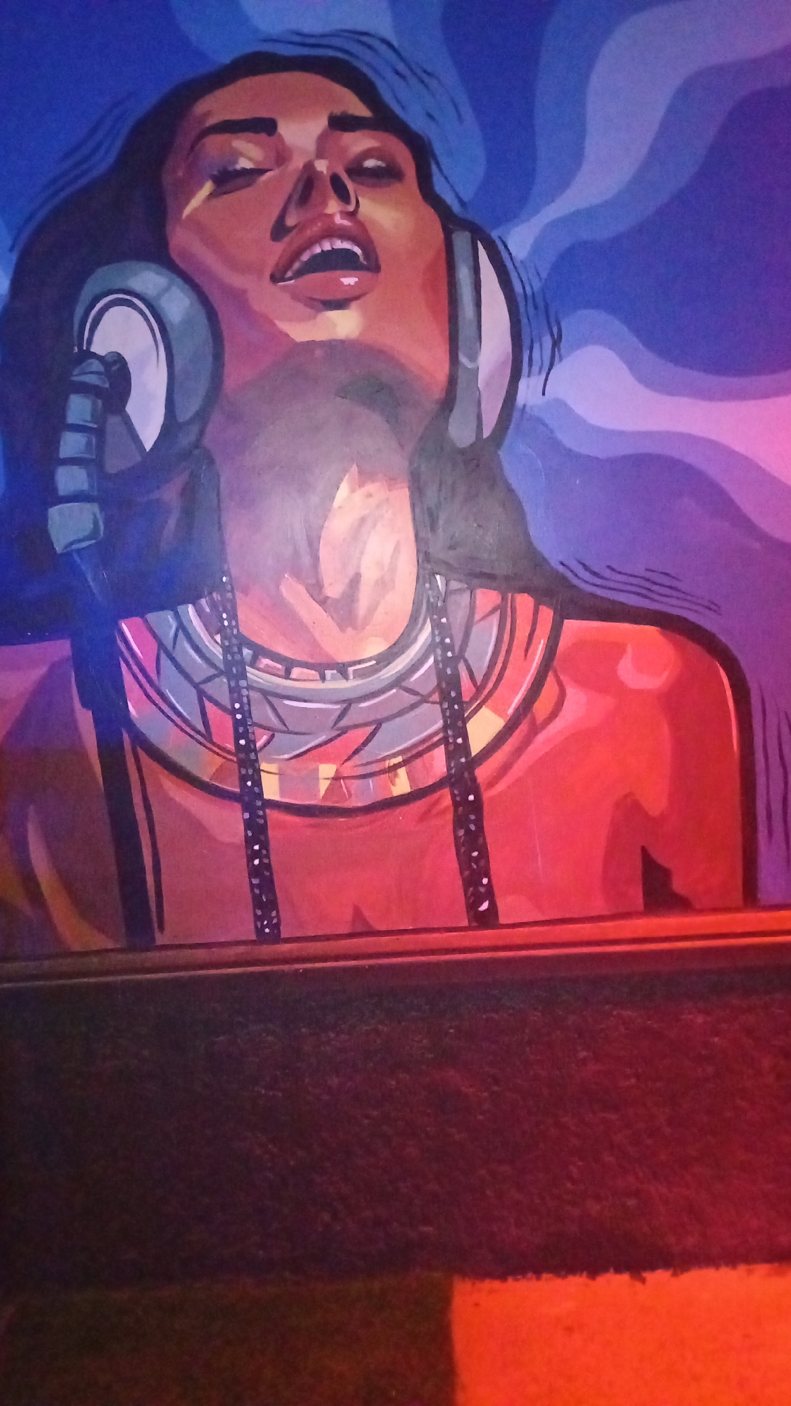 This is a photograph of a mural depicting a woman wearing headphones and a large, ornate necklace. The woman's head is tilted back, and her mouth is open. Swirling lines of blue and purple emanate from behind her head, suggesting sound waves or energetic vibrations. The mural is painted in bold, vibrant colors with a stylized aesthetic. The lower portion of the image shows a dark, textured surface, possibly a stage or seating area, with warm orange light illuminating the bottom edge. There is no information in the image to indicate the location is Howayi, Nigeria.