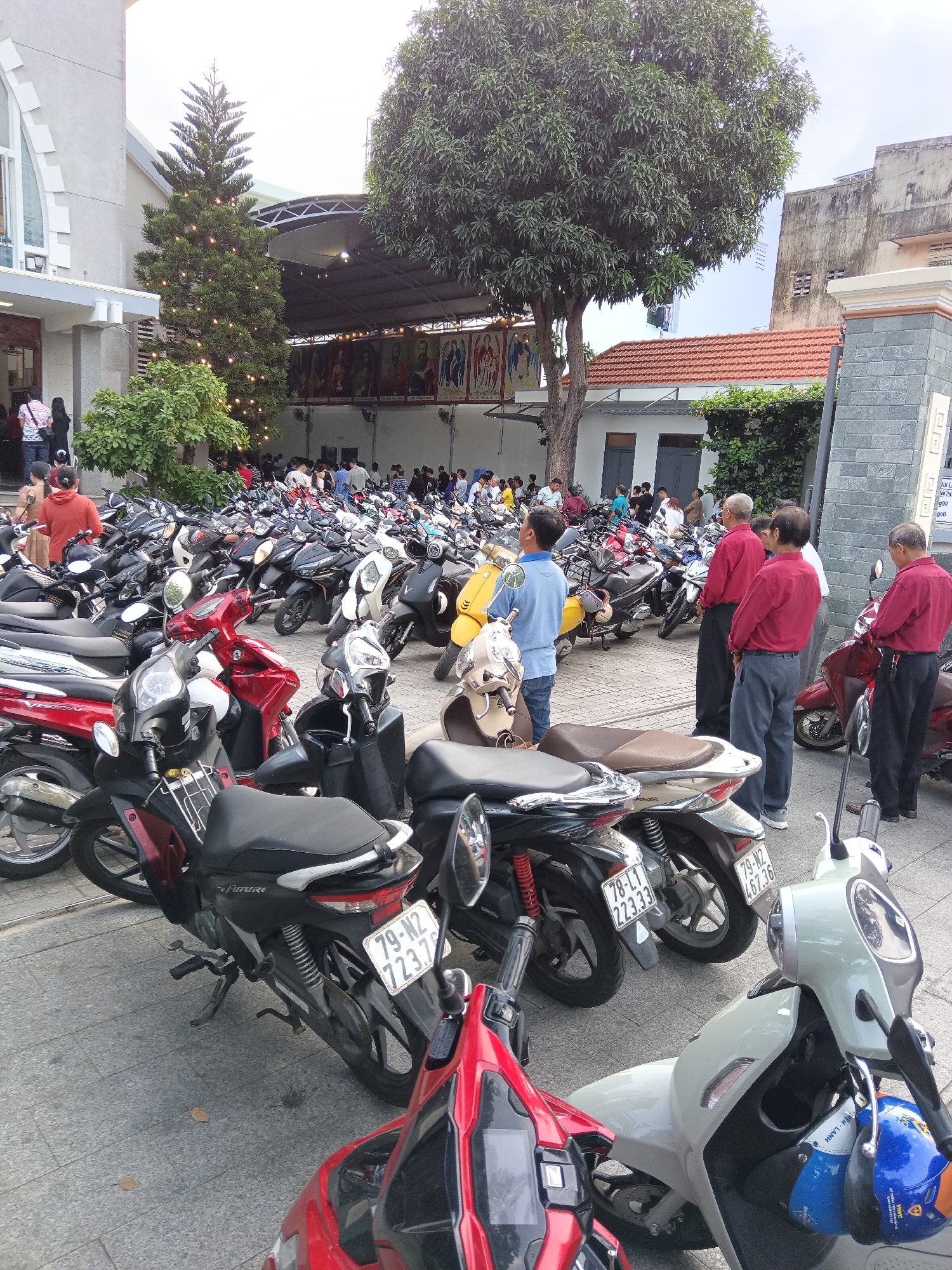This is a street-level, eye-level shot of a large outdoor parking area filled with motorbikes in front of a building that appears to be a church or religious institution in Nha Trang, Vietnam. A tall evergreen tree decorated with lights is prominent to the left of the frame, suggesting a holiday or festive occasion. The building has a light-colored facade with a large arched window and an entrance.
A significant number of people are present, many of them milling about in front of the building or standing near their motorbikes. The crowd appears to be predominantly Asian. Several individuals are wearing casual attire, while a few men in red shirts and dark pants are standing together in the mid-ground, facing away from the camera. A man in a blue shirt is also visible.
The parking area is densely packed with motorbikes of various colors, including red, black, white, and beige. Some motorbikes have visible license plates, such as "79-N2 723.76" and "78-LT 223.33".
The ground is paved with light-colored tiles. In the background, other buildings and trees are visible, along with a large canopy structure extending over part of the area. The overall impression is a busy scene of a community gathering at a religious site, with a significant number of attendees arriving or departing by motorbike.