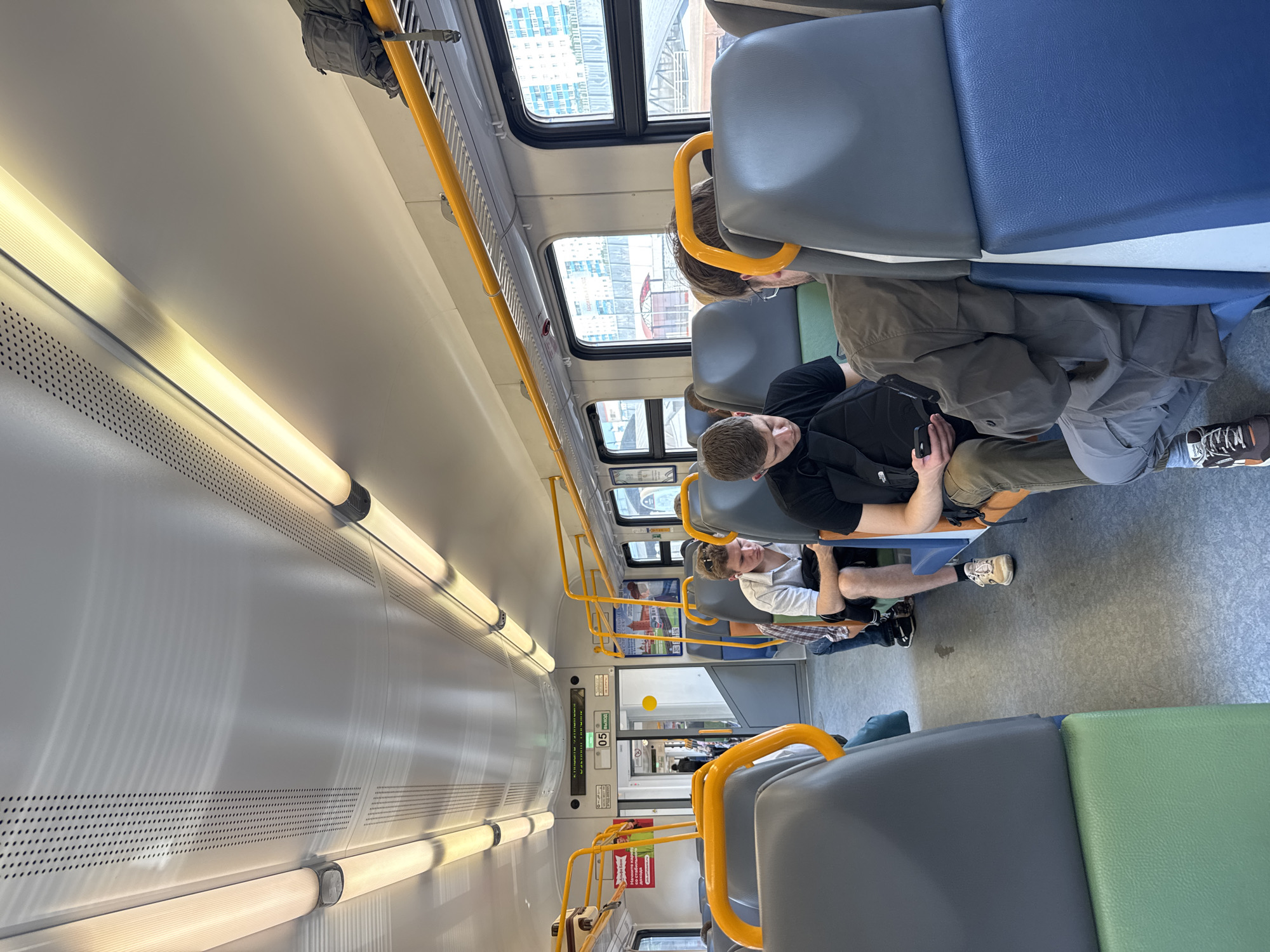 The image shows the interior of a train carriage, likely in Moscow, Russia, based on the Cyrillic text visible on signage. Several passengers are seated, some looking at their phones and others appearing lost in thought. The seats are upholstered in a gray and blue fabric, with yellow grab bars running along the ceiling. The lighting is provided by long fluorescent tubes. The train appears to be moving, as indicated by the blurred view of buildings outside the windows. The overall atmosphere is one of everyday travel.