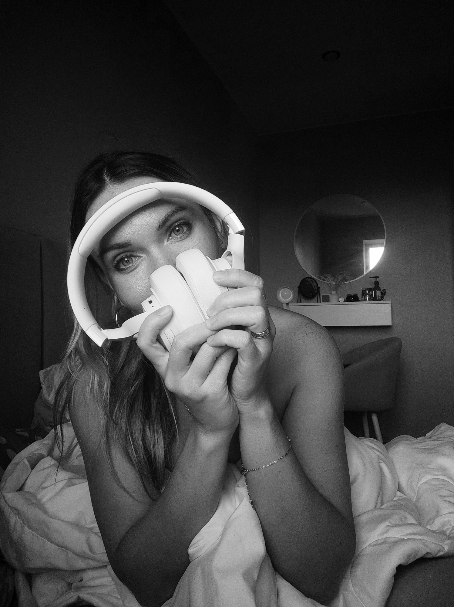 A grayscale close-up photograph depicts a woman in bed, holding white headphones in front of her face. Her eyes are visible above the headphones. She is partially covered by a white duvet. A bracelet is visible on her left wrist, and a ring is on her left ring finger. In the background is a bedroom setting with a round mirror hanging above a shelf, which contains various toiletry-type items, and a chair. The location, Votkinsk, Russia, is not evident from the image.