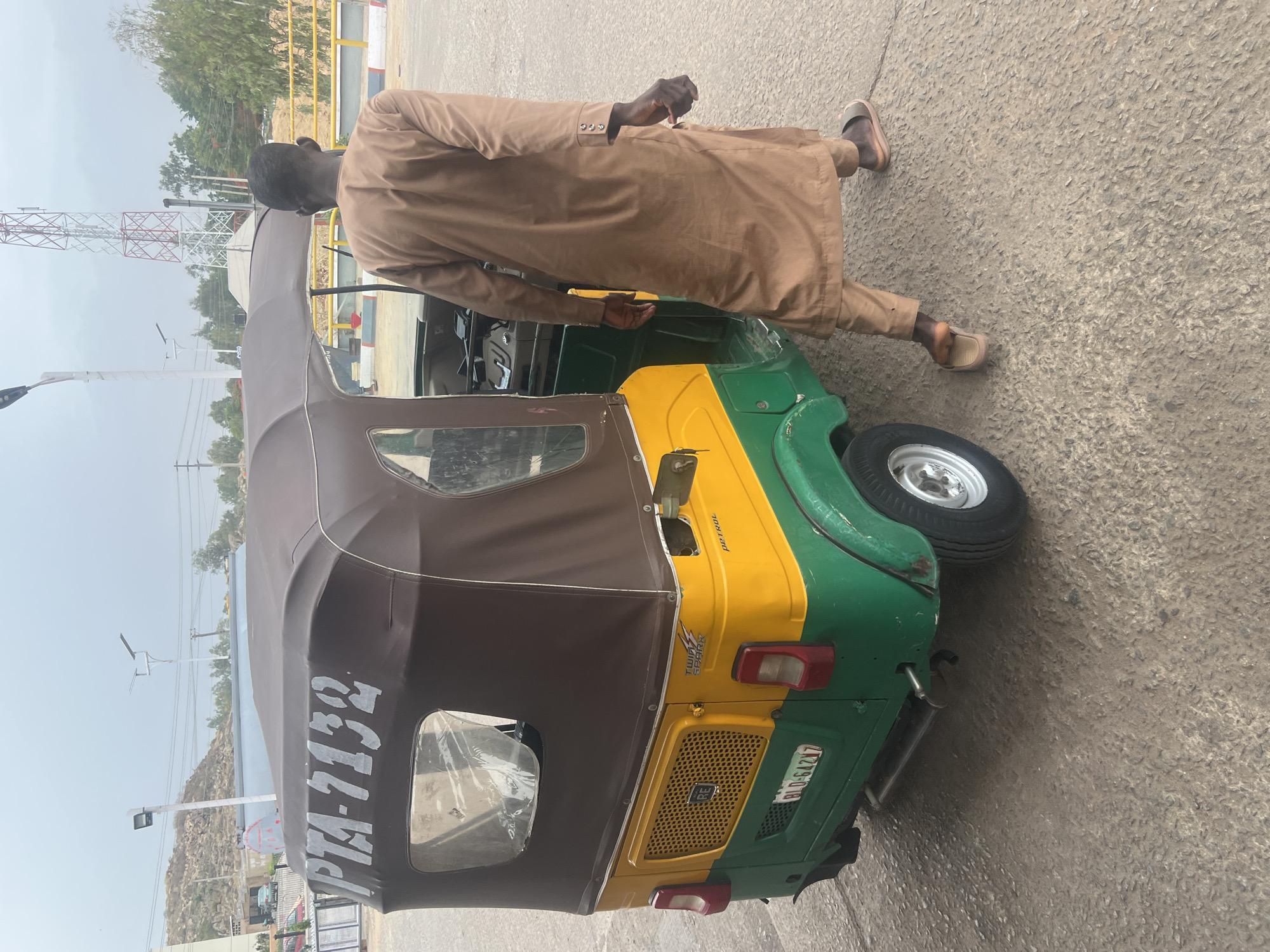 A man wearing a tan traditional outfit stands near a green and yellow tricycle taxi on an asphalt surface. The vehicle displays "PTA-7132" and "BLO-642427." In the background, there is an antenna, a street light, and green vegetation. The setting is Gura Top, Nigeria.