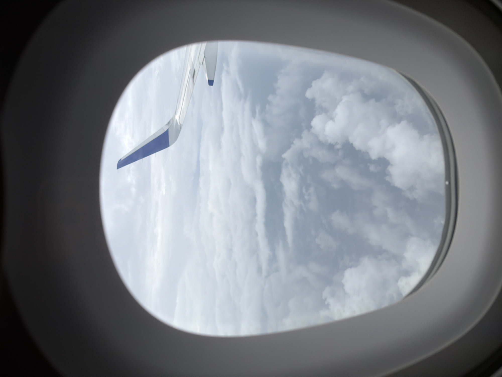 The image shows an airplane wing with a blue tip visible through an oval-shaped airplane window. Below the wing is a vast expanse of clouds. The exterior of the window frame is dark and out of focus, with the interior of the frame appearing as a light gray oval. The scene suggests an aerial view from an airplane. The provided location information, "Lakhiwali, India," is not visually verifiable from the image itself, as the image only depicts clouds and an airplane wing.