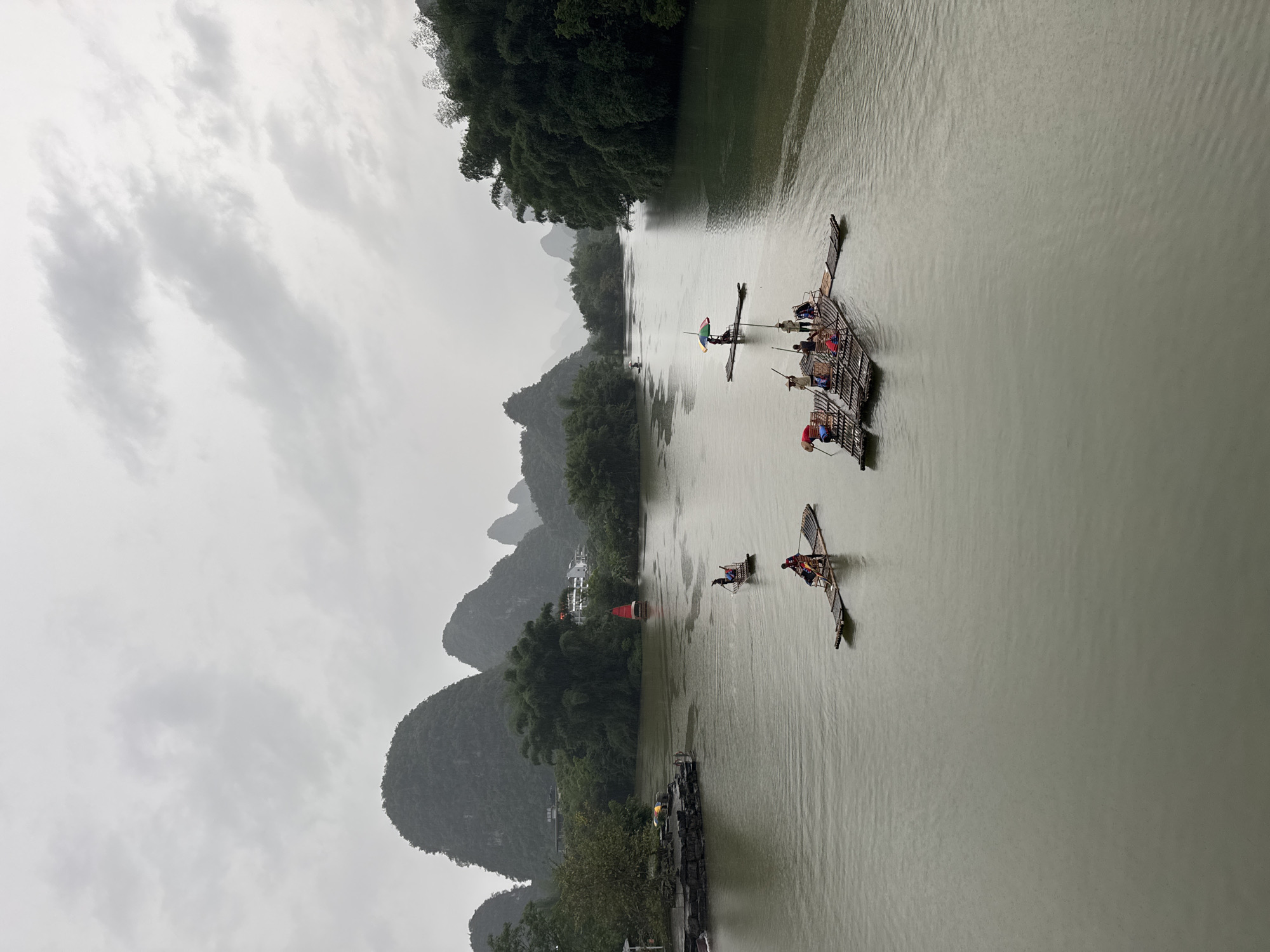 The image captures a scene on the Li River in Guilin, China, under a cloudy sky. The river is flanked by karst mountains covered in lush greenery. Several bamboo rafts are visible on the river, carrying passengers. One raft is larger and holds multiple people, while the others appear to be smaller, carrying fewer individuals. The people on the rafts are observing the scenery and engaging with the boatmen. The river water is a murky grey-green color.