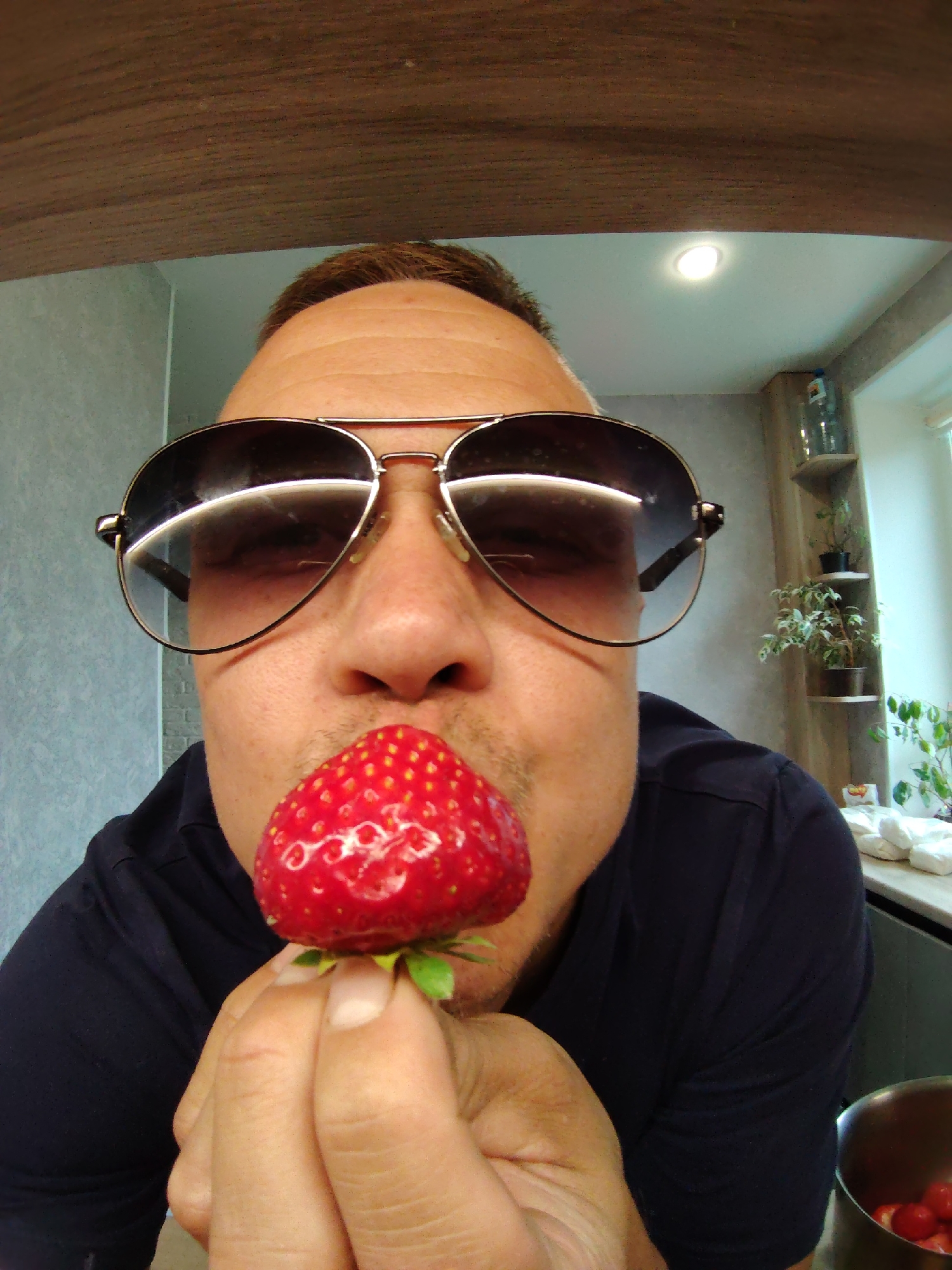The image is a close-up, selfie-style shot of a person wearing aviator sunglasses, holding a strawberry to their lips. The person is wearing a dark blue collared shirt. The background appears to be an indoor kitchen environment, with a wooden beam across the top of the frame, a light fixture on the ceiling, and shelves with plants and bottles visible in the background. There is also a metal pot with some red objects, possibly more strawberries, in the lower right corner. The overall lighting suggests an indoor setting, possibly during the daytime or with artificial lighting. No specific location within Chaikovsky, Russia is discernible from the image.