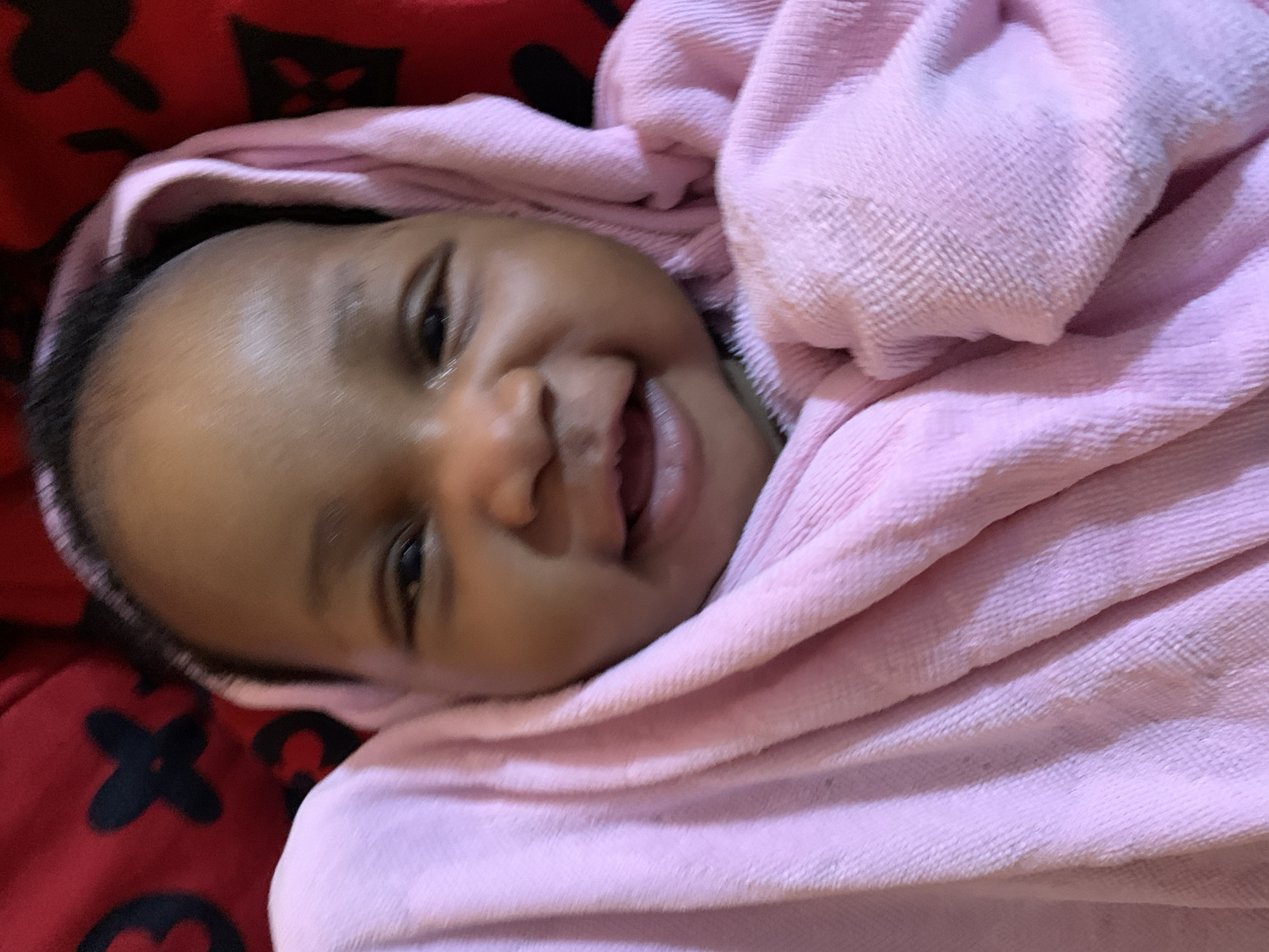 A close-up shot depicts a smiling infant wrapped in a pink blanket, positioned on a red fabric with a black repeating floral or quatrefoil pattern. The baby's face is the primary focus, with visible dark eyes, a small nose, and an open mouth revealing the tongue and gums. The pink blanket is loosely draped around the baby, partially obscuring the background. The image suggests a cozy and intimate setting. The information is insufficient to make any statement of location.