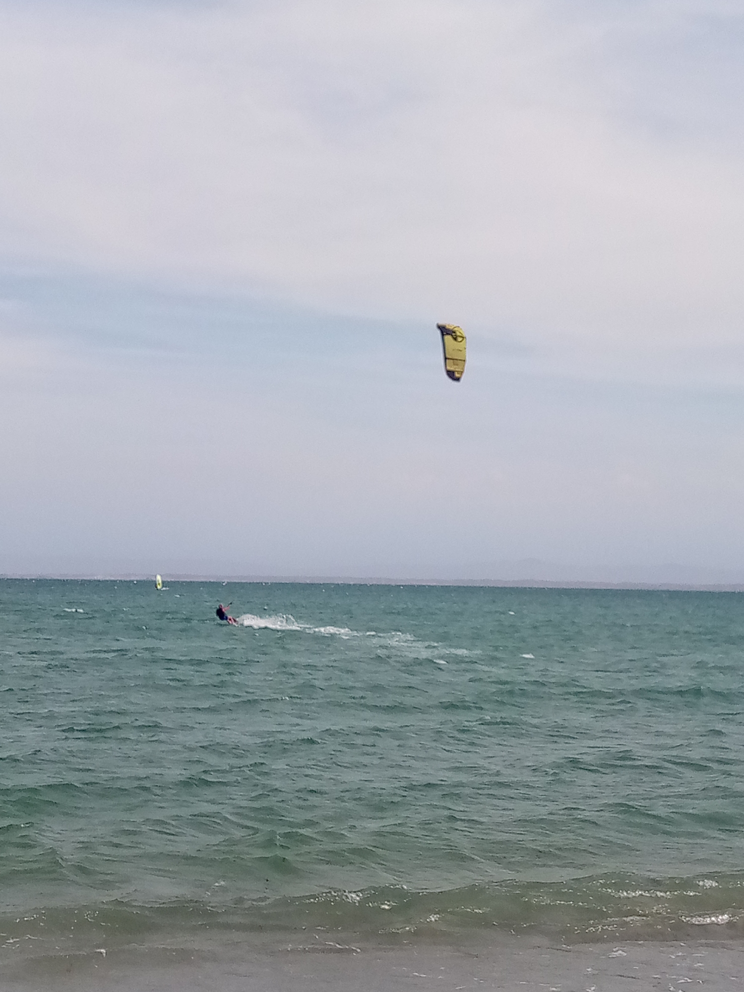 The image shows a kiteboarder in the sea off El Yaque, Venezuela. The kiteboarder is moving from left to right. A yellow kite is in the air above the kiteboarder. A windsurfer is visible in the distance to the left of the kiteboarder. The water is choppy. The sky is overcast. The shoreline is visible in the foreground.