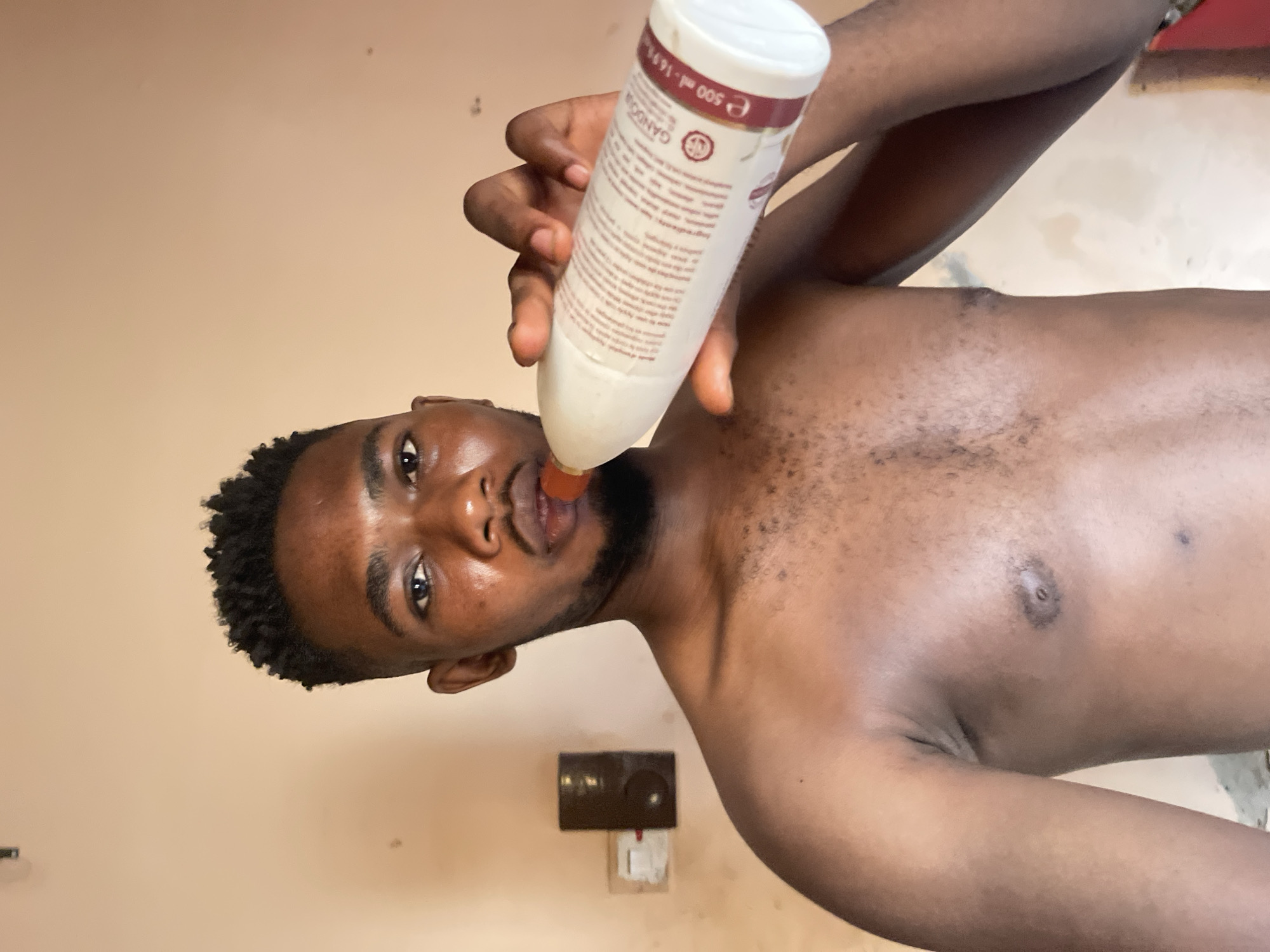 A shirtless, dark-skinned male with short, curly hair, a beard, and facial hair around his chest is holding a white bottle with red lettering and markings up to his mouth. The bottle is tilted downwards. The background includes a light beige wall with an electrical outlet. The location is in Muchiya, Nigeria, based on the implied geographic context of the prompt.