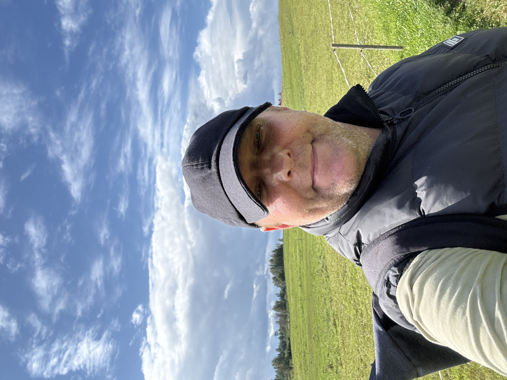 A selfie taken outdoors shows a man wearing a black baseball cap, a black puffer vest over a tan long-sleeved shirt, and a backpack. He is smiling and looking at the camera. The background consists of a green grassy field with a few trees in the distance and a cloudy blue sky above. A wire fence is visible on the right side of the frame. The location is Putiniškės, Lithuania.