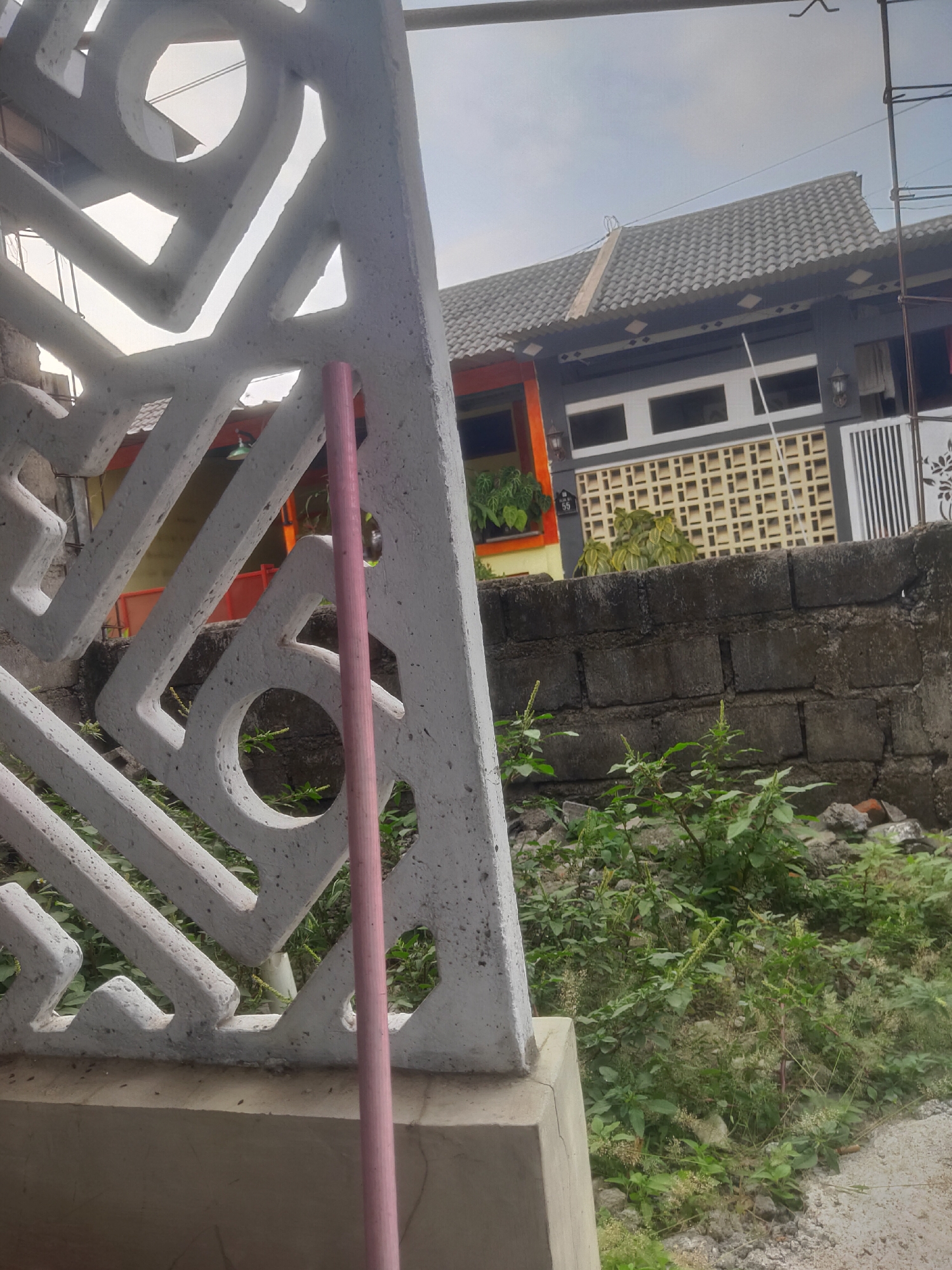 The image depicts a street view with a focus on a decorative concrete fence in the foreground. This fence has an abstract geometric design with various cutouts. A pink cylindrical object, possibly a pole or a pipe, is leaning against the fence. Behind the fence, a low brick wall is visible, overgrown with green vegetation. Further in the background, a house with a tiled roof and dark gray walls is seen. The house features white window frames and decorative lattice work. The sky is overcast. The scene appears to be an outdoor environment, likely in a residential area.