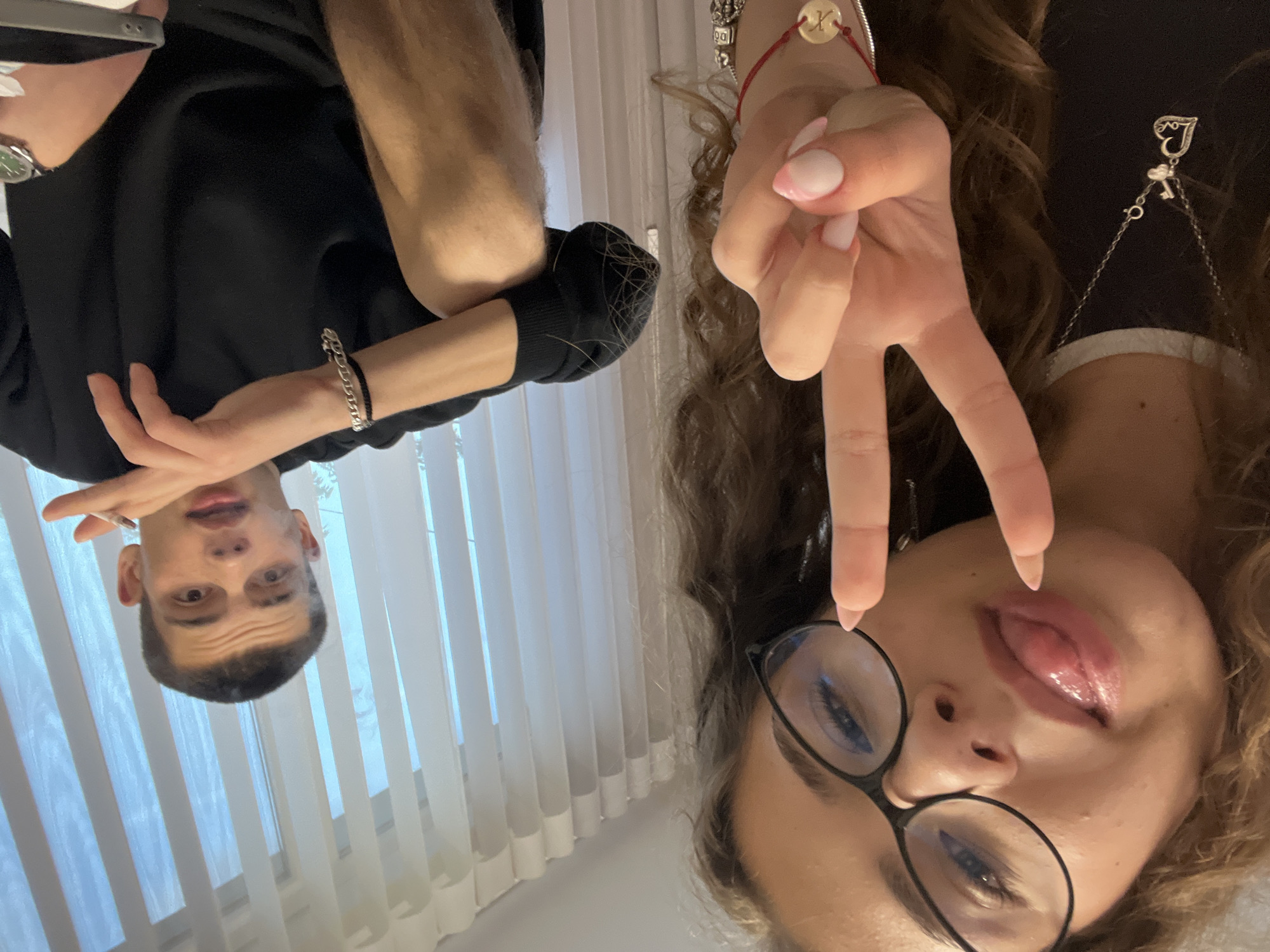 Two people are in a vertically oriented selfie image. One person is male, fair-skinned, with short dark hair, dark eyebrows, brown eyes, and is wearing a black top. He is positioned on the left, looking at the camera. The other person is female, fair-skinned, with brown curly hair, wearing glasses and a dark top with a white collar. She is making a peace sign with her right hand near her face and sticking out her tongue. In the background, there are vertical blinds. The location, Karavelovo, Bulgaria, is not discernable from the image.