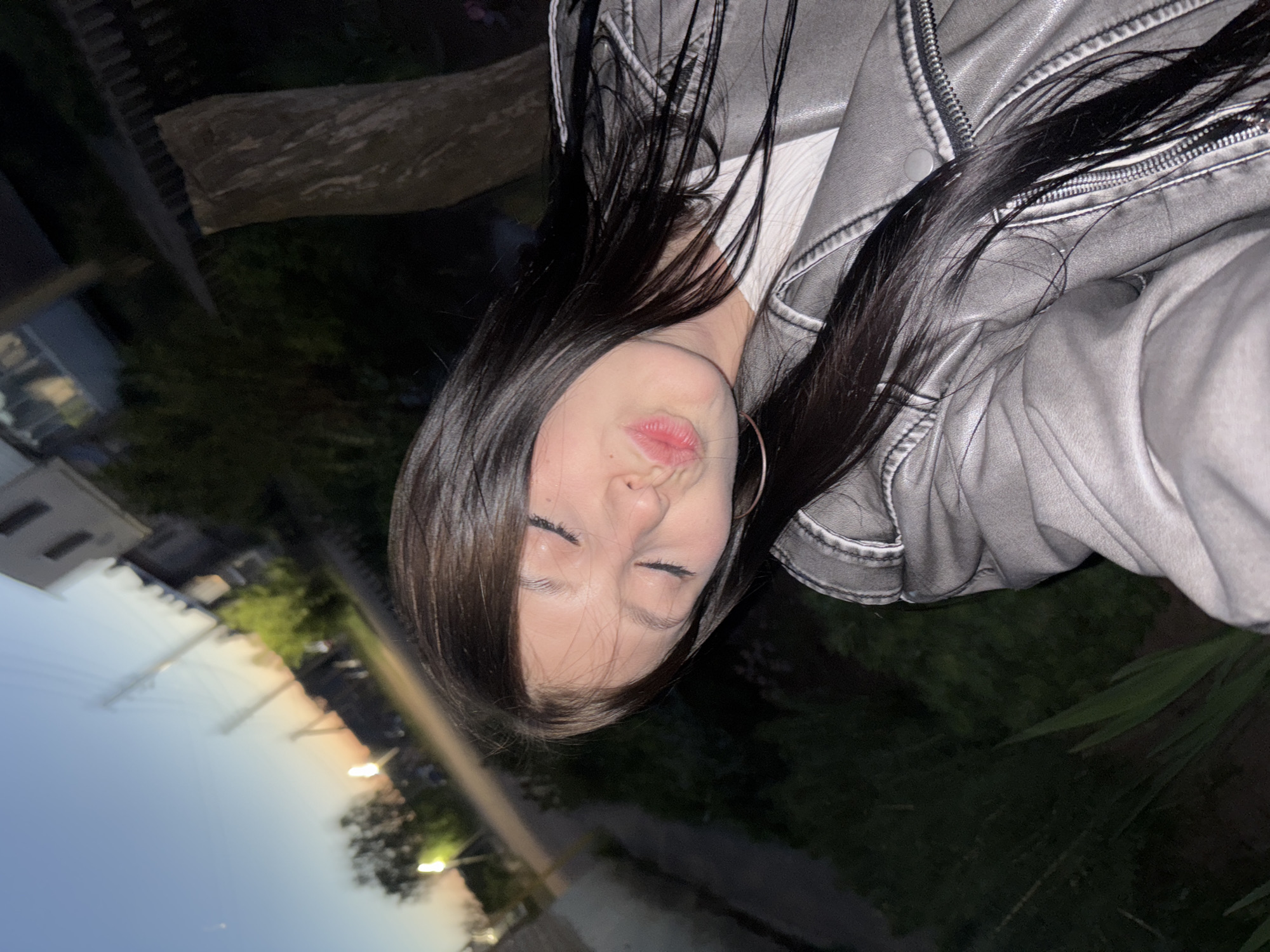 A woman with long, dark hair is taking a selfie. She is wearing a light grey leather jacket over a white shirt. Her eyes are closed and her lips are pursed. An earring is visible on her left ear. The background shows a twilight sky and some greenery, with glimpses of buildings and streetlights. The location appears to be an outdoor area in Фоки, Russia.