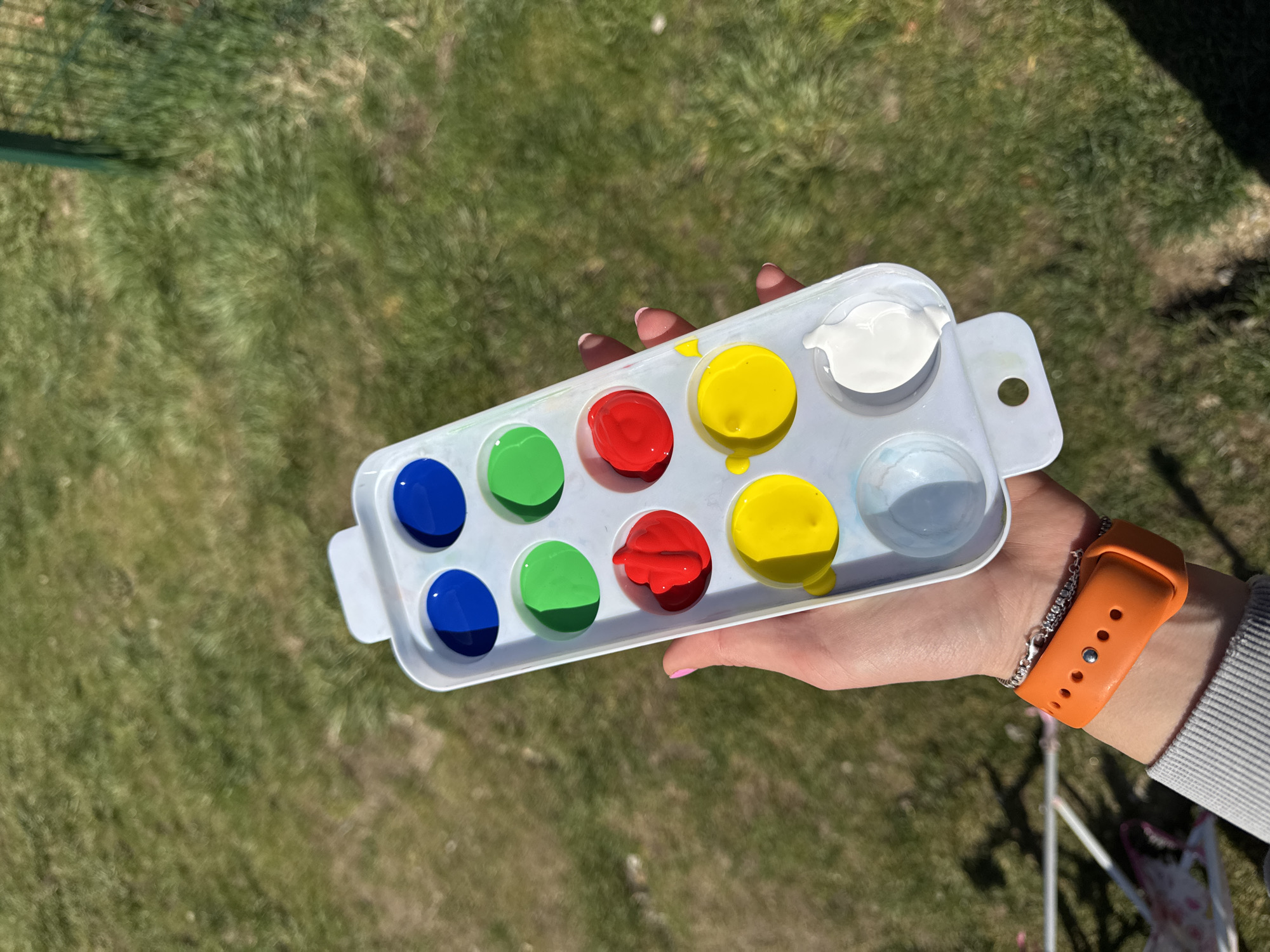 The image shows a person holding a white plastic paint palette in their left hand, which contains dabs of blue, green, red, yellow, white, and light blue paint. The person is wearing an orange watch on their left wrist. The background is a blurred expanse of green grass. The location is not discernible from the provided image and cannot be determined to be in Sofia, Bulgaria.