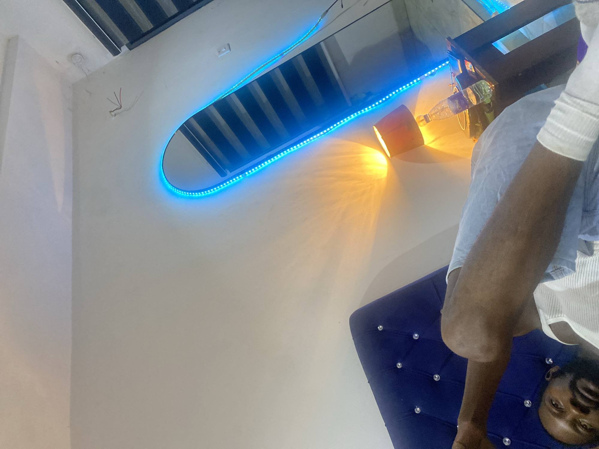 A rotated view of an interior scene shows a person sitting on a blue tufted headboard. The wall has a rectangular opening illuminated by blue LED lights. An orange lamp emits a warm light. The person is wearing a white tank top and light blue denim shorts. Location: Abuja, Nigeria.