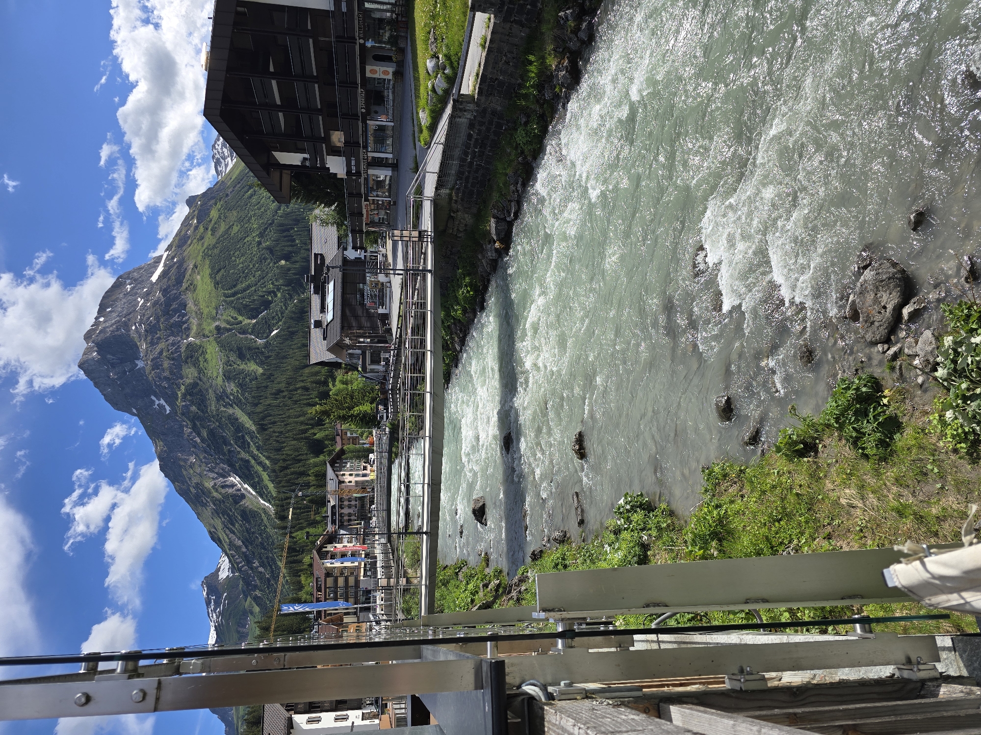 The image depicts a scenic view of the Lech river flowing through the village of Lech, Austria. In the foreground, a modern metal and wooden pedestrian bridge spans the turbulent, greenish-blue water, which is characterized by white rapids and scattered rocks. The riverbanks are lined with lush green grass and vegetation, with more rocks visible in the shallow areas.
Beyond the river, the village of Lech unfolds. Buildings with traditional Alpine architecture, featuring pitched roofs and wooden balconies, are visible. Several shops and businesses with signage such as "SPORTALP," "SKI SCHULE LECH," and "WALDHART" are apparent. A prominent dark-colored building with multiple stories and balconies stands to the right.
In the background, the majestic Arlberg mountain range dominates the skyline. The mountains are steep and rugged, with patches of green grassland and dense coniferous forests covering their lower slopes. Snow can be seen on the higher peaks, suggesting a season where winter activities are concluding or have recently ended. The sky is a vibrant blue, dotted with fluffy white clouds. The overall scene conveys a peaceful and picturesque mountain village atmosphere.