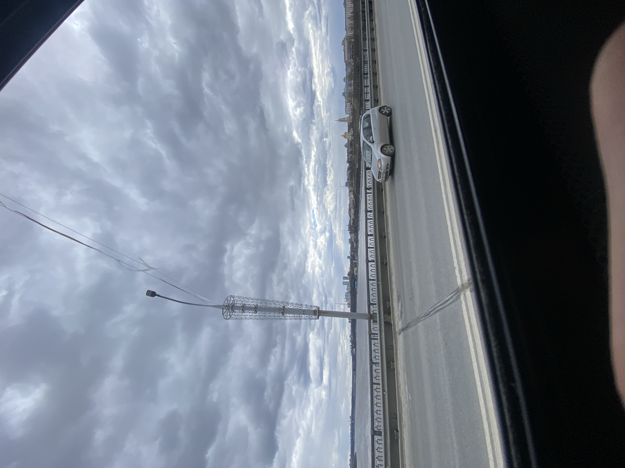 The image captures a view from inside a vehicle, possibly a car, looking out onto a bridge in St. Petersburg, Russia. The street lamp with its cage-like structure is a prominent feature, and a silver car is traveling along the bridge. The Neva river can be seen to the side of the bridge, and the skyline of St. Petersburg is visible in the distance. The sky is overcast with heavy clouds.
