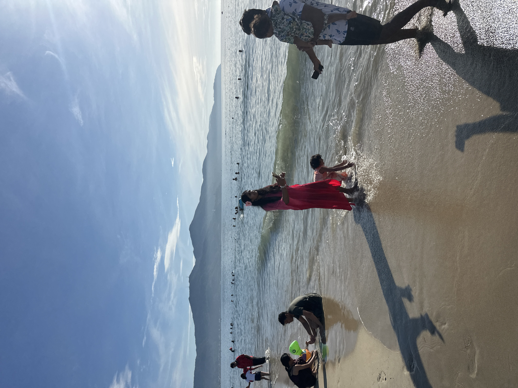 The image depicts a sunny day at a beach in Đà Nẵng, Vietnam, likely Mỹ Khê Beach given its popularity and characteristic mountain backdrop. The scene is populated with numerous people enjoying the ocean and the sandy shore. In the foreground, a family is visible on the beach. A man on the right is holding a baby and looking at his phone. To the left, two individuals are sitting in the shallow water, with a child playing with a green toy in front of them. Further out in the ocean, many people are swimming and wading. The background features a coastline with lush green mountains under a bright blue sky with scattered white clouds. The sun appears to be setting or rising, casting a warm glow and long shadows on the sand.