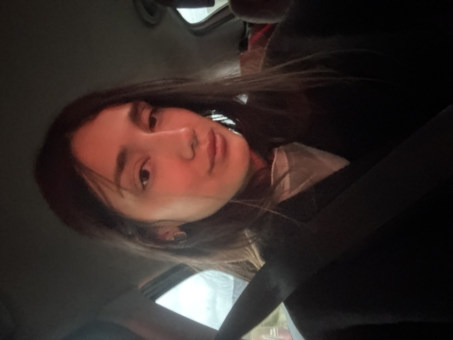 A person with long dark hair is sitting in a car, wearing a seatbelt. They are looking directly at the camera with a slight smile. The background is partially visible through the car windows and is out of focus. The interior of the car is dark, with the seatbelt prominently displayed across the frame. The lighting creates shadows on the person's face.
