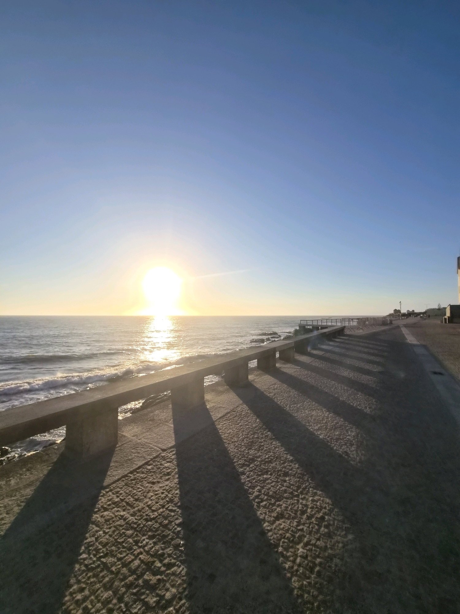 A wide, angled, full shot captures a scenic oceanfront promenade in Esposende, Portugal, during a bright, clear day. The sun, low on the horizon, casts a brilliant golden glow across the tranquil sea, reflecting a shimmering path of light on its surface. The sea itself is calm, with small waves gently lapping at the shore.
In the foreground, a concrete promenade runs parallel to the ocean. A low, concrete railing with square pillars borders the walkway, separating it from the sea. The promenade is paved with rough, textured stones that are illuminated by long, distinct shadows cast by the railing. The shadows stretch diagonally across the paved surface, creating a visually striking pattern.
To the right of the promenade, a paved walkway extends further into the distance, lined with metal barricades. Further along the promenade, several low-rise buildings are visible, with one particularly tall, light-colored structure to the far right of the frame. The sky above is a clear, vibrant blue, gradually fading to a paler hue near the horizon. No people are visible in the frame. The overall impression is one of peaceful, early morning or late afternoon serenity by the sea.
