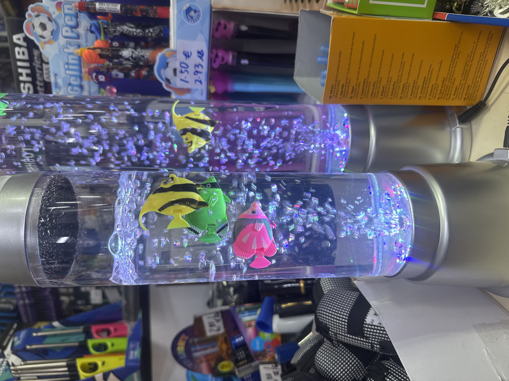 The image displays a close-up view of two bubble lamps with artificial fish, set against a background of store merchandise. The lamps are cylindrical and filled with water, illuminated by blue and purple lights at the base, causing tiny bubbles to rise. In the left lamp, a pink and green artificial fish are visible. In the right lamp, a yellow and black artificial fish is prominent.
To the right of the lamps, a display of pens is visible, some with colorful caps. Above the pens, a sign reads "SHIBA Batteries AAA" and "Gelink Pen" with a soccer ball motif. Below this, a price tag indicates "1.50 €" and "2.93 лв", the latter being the Bulgarian lev. The overall environment suggests a retail setting, likely a stationery or gift shop.
A white box filled with black and white patterned items, possibly socks or fabric rolls, is located to the left of the bubble lamps. In the far left background, a display of lighter-like objects in various bright colors (yellow, green, pink) is partially visible.
The presence of the Bulgarian currency "лв" strongly indicates the location is within Bulgaria. Given the general appearance and the type of merchandise, a plausible location in Sofia would be a store within a shopping mall or a dedicated stationery and gift shop. The specific store or street cannot be identified from the image alone. There are no people visible in the frame.