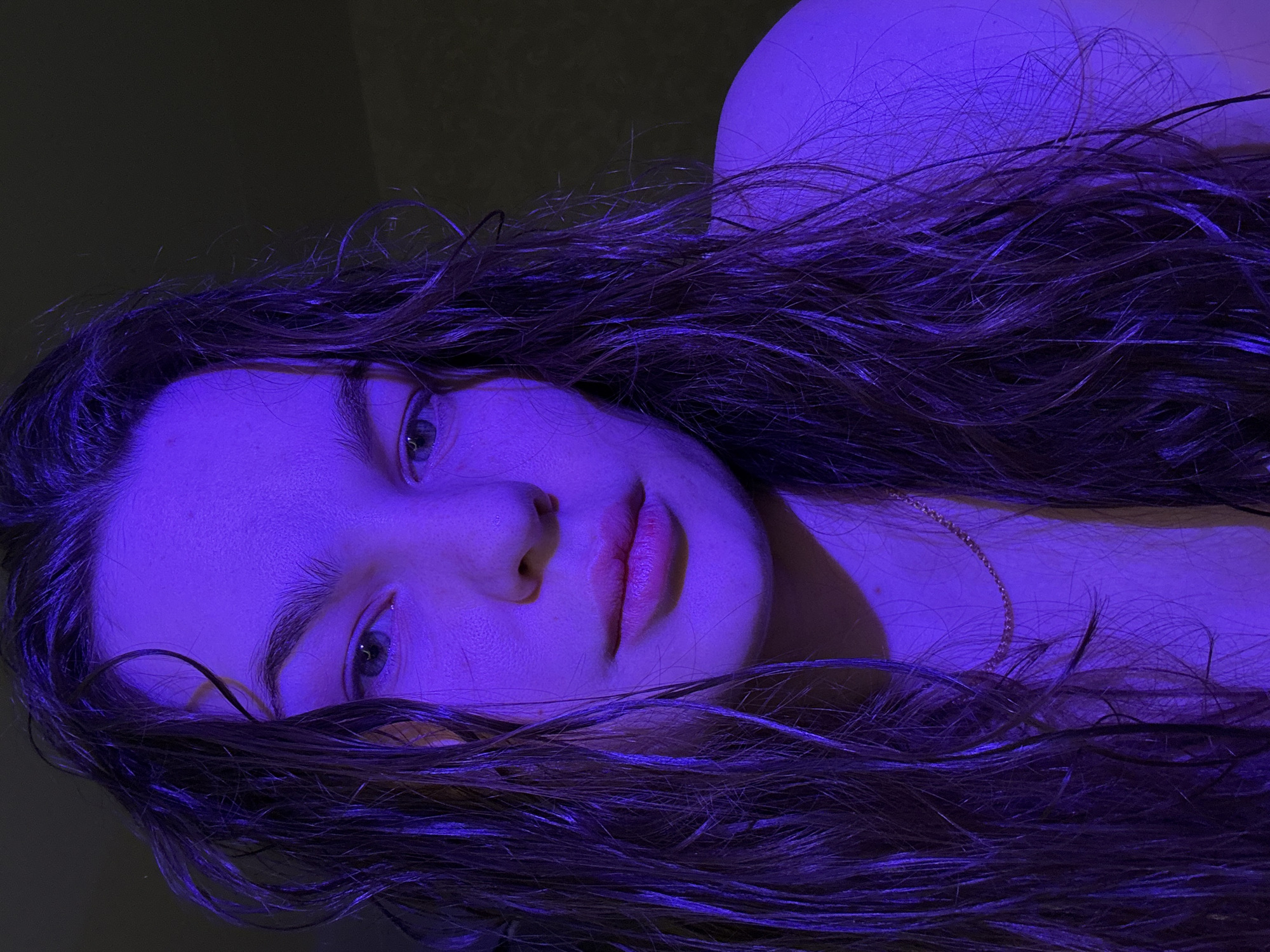 A close-up portrait of a young woman with wet, wavy brown hair, illuminated by a strong blue light source. Her face is angled towards the camera, and her eyes are open. She is wearing a delicate gold necklace. The background is dark and indistinct. The location in Chaikovsky, Russia cannot be determined from this image.