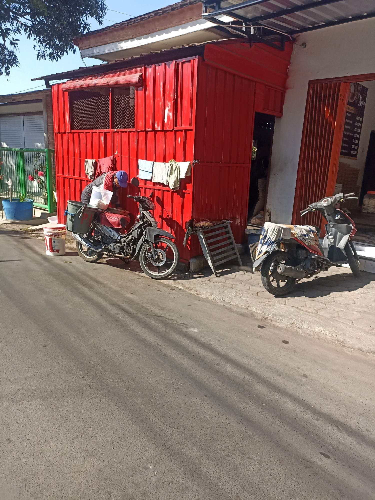 This is a forensic, matter-of-fact description of a street scene in Mojopanggung, Indonesia.
The scene is set on a paved street with a sidewalk made of interlocking hexagonal stones. A bright red, corrugated metal structure, likely a small shop or kiosk, dominates the right side of the image. A window with a bamboo blind is visible on the left side of this structure, and laundry hangs on a line attached to it. Two motorcycles are parked on the sidewalk in front of the red structure. The motorcycle on the left is black and grey, with a person wearing a blue cap and red jacket leaning over it, interacting with a white plastic container. A large white and red bucket is next to this motorcycle. The motorcycle on the right is black and orange, covered with a patterned cloth. A metal rack with straw is leaning against the red structure to the left of the second motorcycle. The entrance to the red structure appears to be an open doorway. Further to the left, a green metal fence encloses a small area with a blue plastic tub and some pink flowers. The background includes more buildings and trees, with a clear blue sky above. The lighting suggests it is daytime, with distinct shadows cast on the ground.