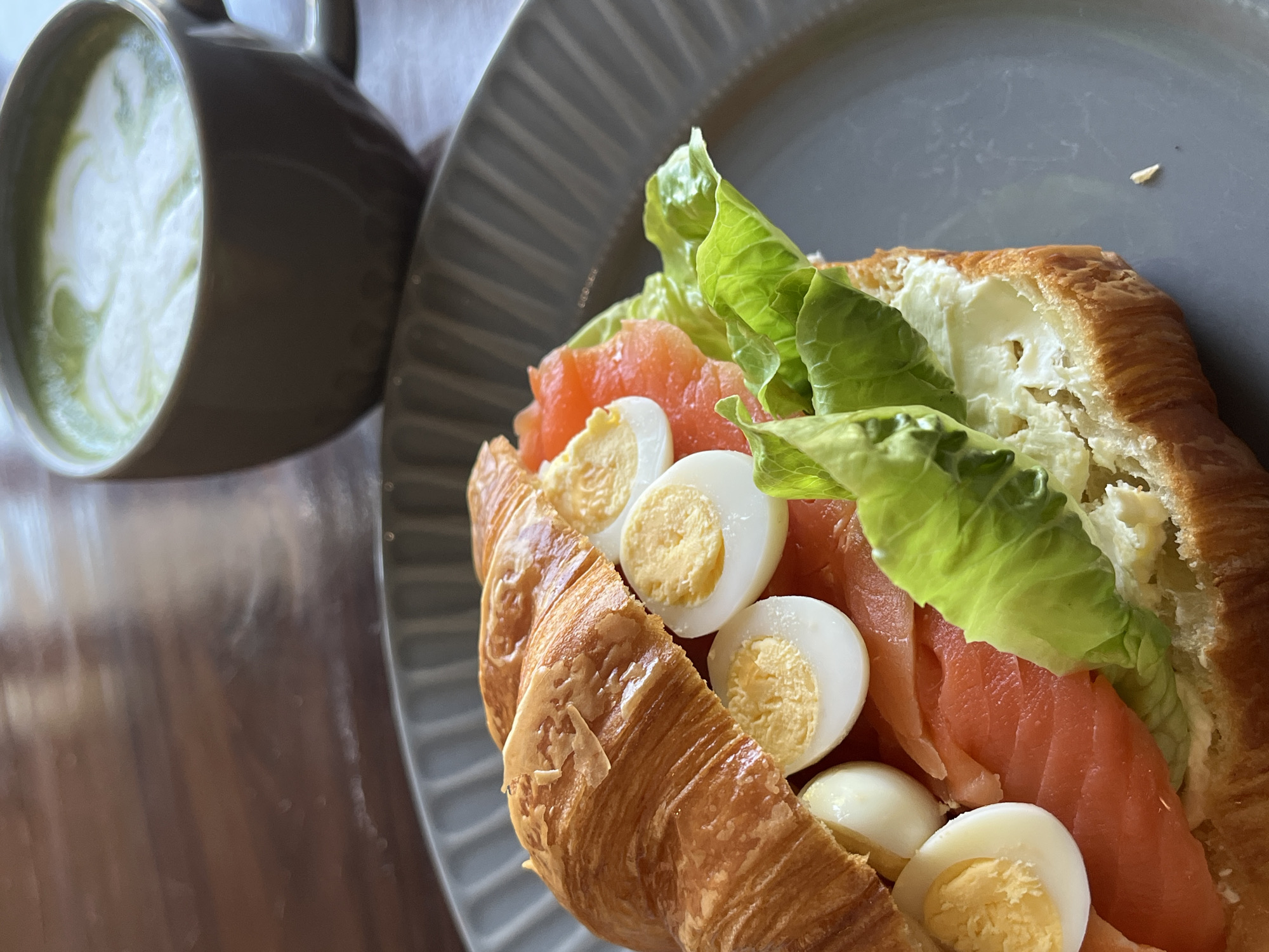 A close-up shot shows a croissant sandwich filled with cream cheese, salmon, lettuce, and halved hard-boiled eggs, on a grey plate. Beside the plate is a grey mug filled with a green and white beverage, possibly a matcha latte. The objects rest on a wooden surface. The location in Kuta Selatan, Indonesia, cannot be determined from the image.
