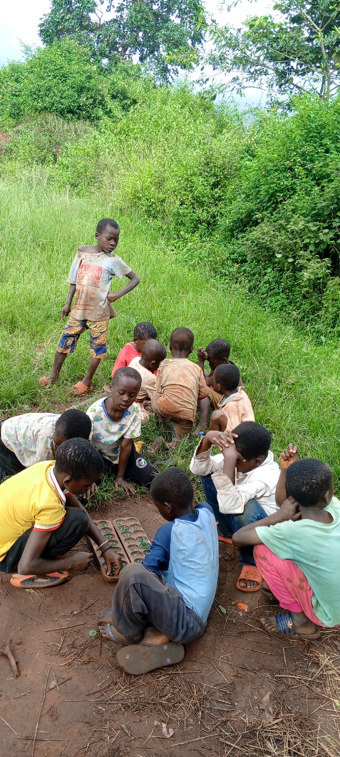 The scene captures approximately ten children, primarily males, gathered outdoors in Ngalula, Democratic Republic of the Congo. The immediate ground is reddish-brown earth with sparse vegetation and debris, transitioning to dense green grass, bushes, and trees in the mid-ground and background under an overcast sky.
A central activity involves two boys seated on the ground, manipulating small green spherical game pieces on a wooden Mancala-style board game. Eight other children are positioned around them, observing the game or seated nearby. One boy stands on the left, holding a thin stick, with hands placed on his hips.
The children, ranging in apparent age from approximately 5 to 12 years, wear varied, often soiled or worn, casual clothing. Footwear includes flip-flops on several individuals, while others are barefoot.