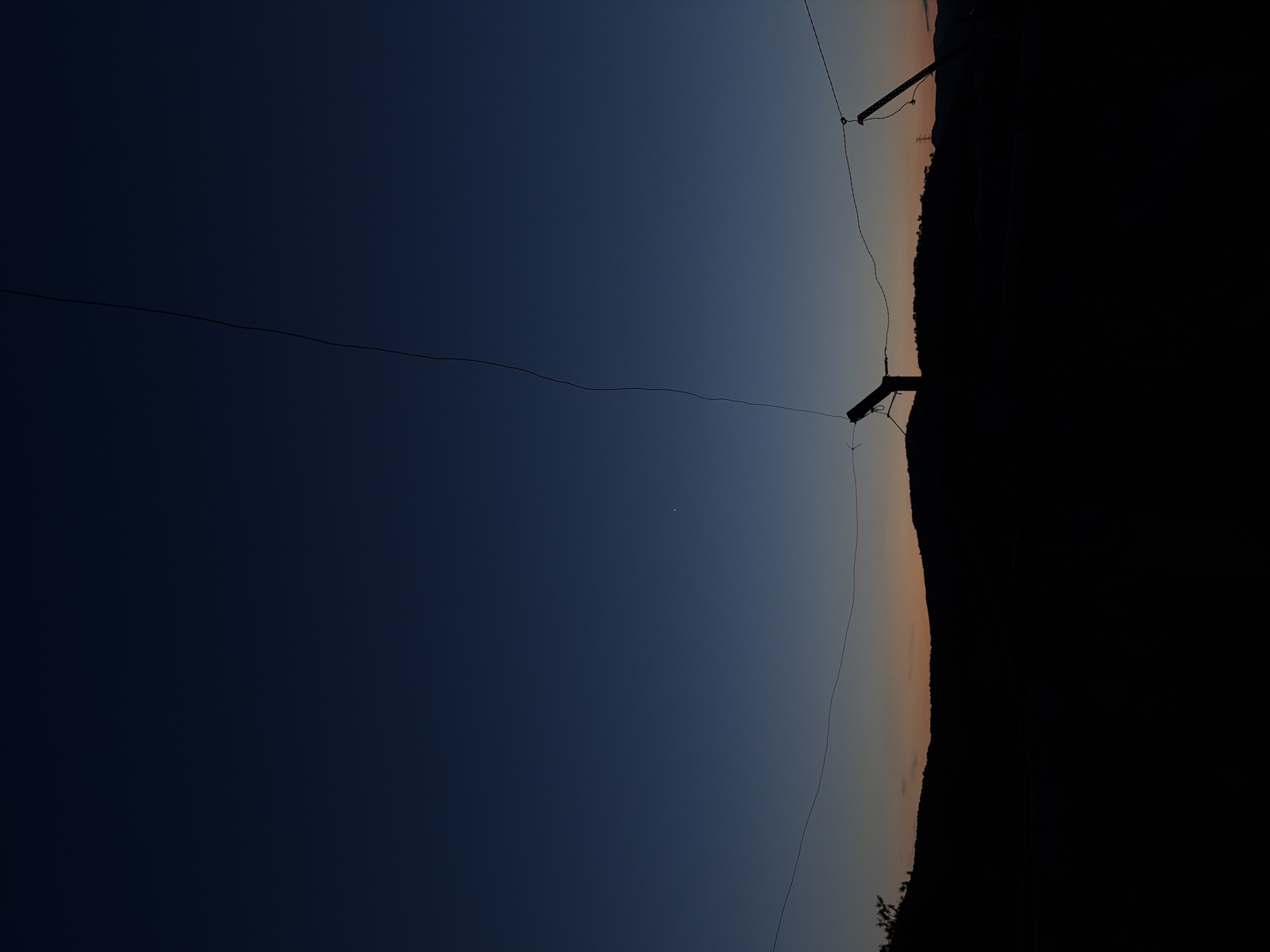 This is a landscape photograph depicting a twilight scene, likely at dusk or dawn, in Sant Martí Sarroca, Spain. The sky dominates the frame, showcasing a striking gradient from a deep, dark blue at the top to a softer, warm orange and pale yellow closer to the horizon. A small, bright white dot, which could be a star or a planet like Venus, is visible in the upper central portion of the dark blue sky.
Stretching horizontally across the middle ground are several thin, dark lines, characteristic of power lines or communication wires. These wires are strung between two utility poles, which appear as dark silhouettes against the sky; one pole is partially visible on the far left, and another on the far right.
The lower part of the image features the dark, undulating silhouette of a landscape, possibly hills or mountains covered in trees, standing out against the lighter, warmer tones of the horizon. The scene evokes a sense of tranquility and the quiet beauty of twilight. No people or specific activities are visible, and the weather appears clear. There is no discernible text in the image.