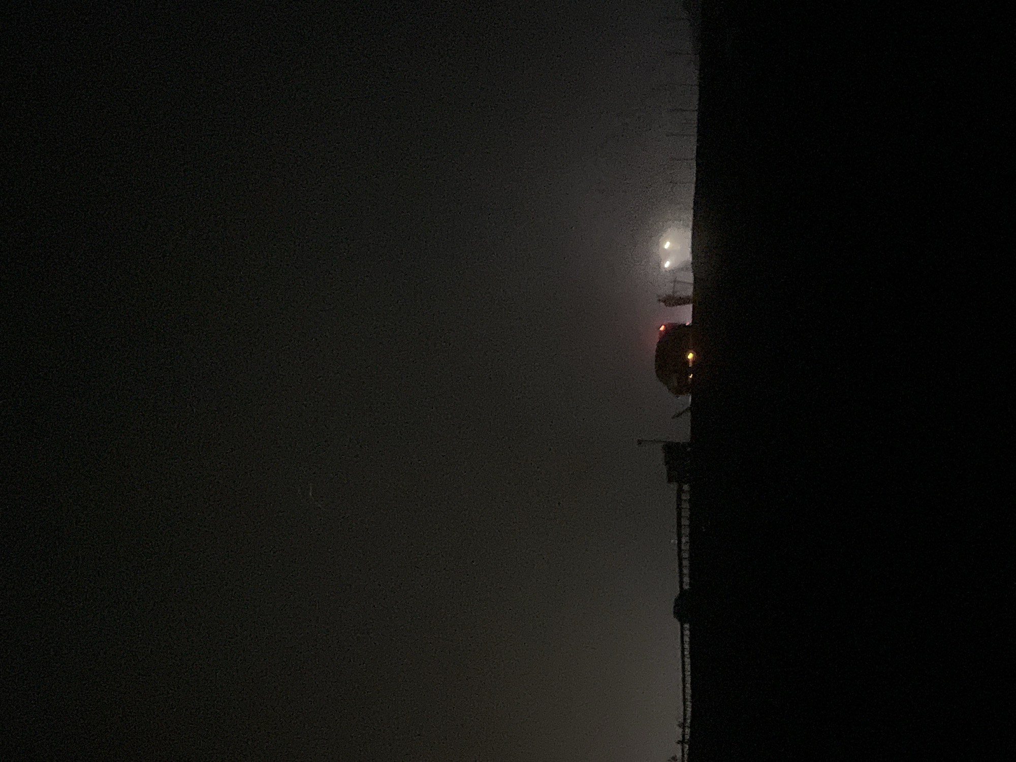 In a dimly lit nighttime scene, the silhouette of a dark horizon is visible against a foggy sky. The scene appears to be a road or highway with lights from vehicles and possibly a structure, suggesting an outdoor urban or semi-urban environment. The lighting is low, creating a sense of mystery and obscurity.