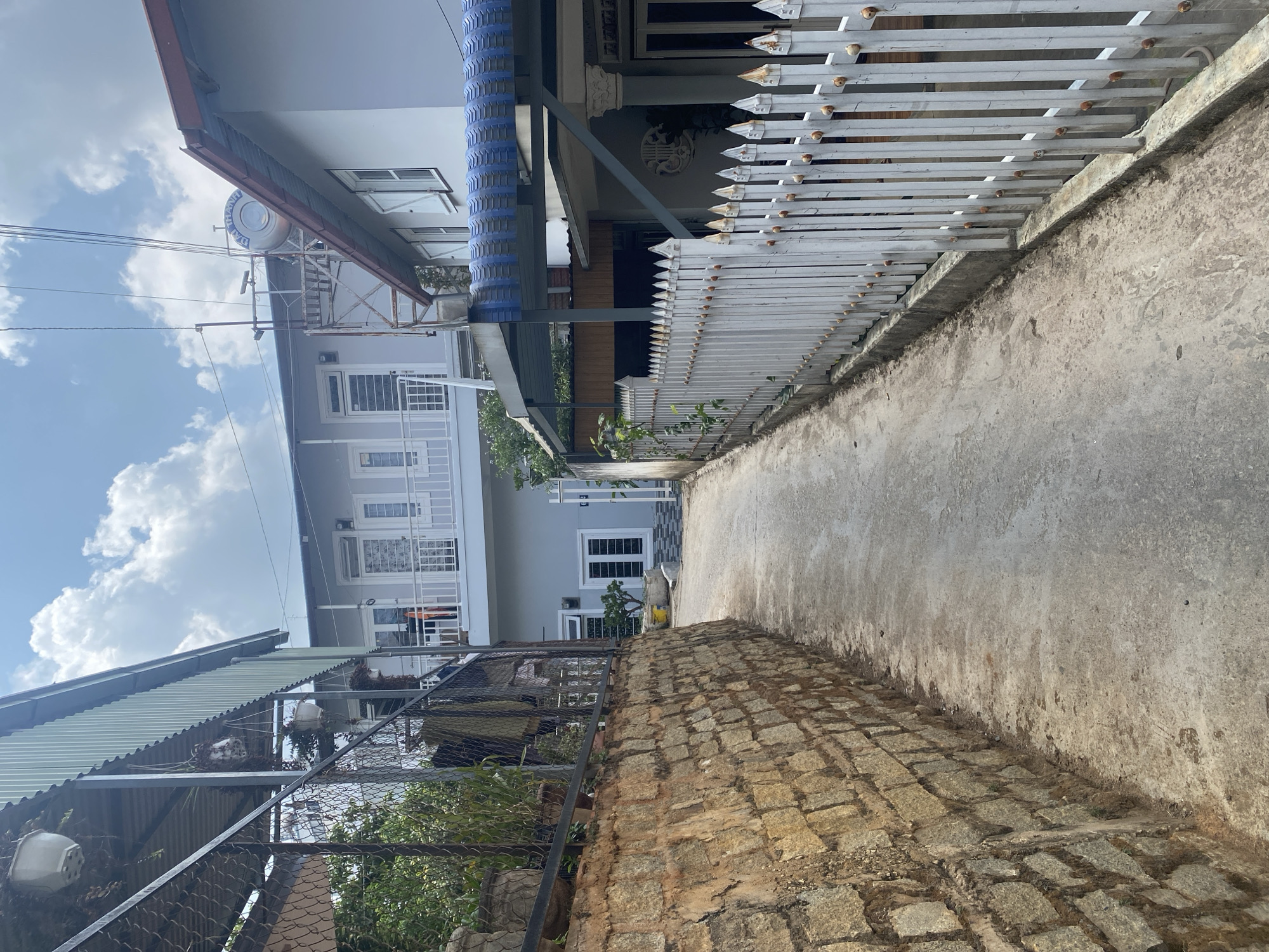 The image depicts a narrow residential alleyway under a bright, partly cloudy sky, likely in Lâm Viên - Đà Lạt, Vietnam. The scene is static with no people or immediate activity.
On the left side of the alley, a weathered retaining wall made of rough-hewn stones or cobblestones forms the base. Above this, a wire fence supports an angled corrugated metal roof, shielding an area with visible plants. Further along, a multi-story house with a light-colored facade features multiple windows and balconies with white railings. A large, light-colored water tank is prominently situated on the roof of this house. Overhead, utility lines traverse the sky.
On the right side, a white picket fence, showing signs of age and some rust on its decorative spikes, runs along a low concrete wall. Behind this fence, parts of a light-colored building are visible, including windows and a distinctive circular wall decoration featuring the traditional Chinese double happiness symbol (囍). Further back, the building has a distinctive blue, possibly corrugated or tiled, roof section.
The alley's surface is a mix of worn concrete and a rougher, earthy path with some stones. The clear blue sky with scattered white clouds indicates a sunny daytime. The water tank on the left house is branded with the text "BÁCH THÀNH".