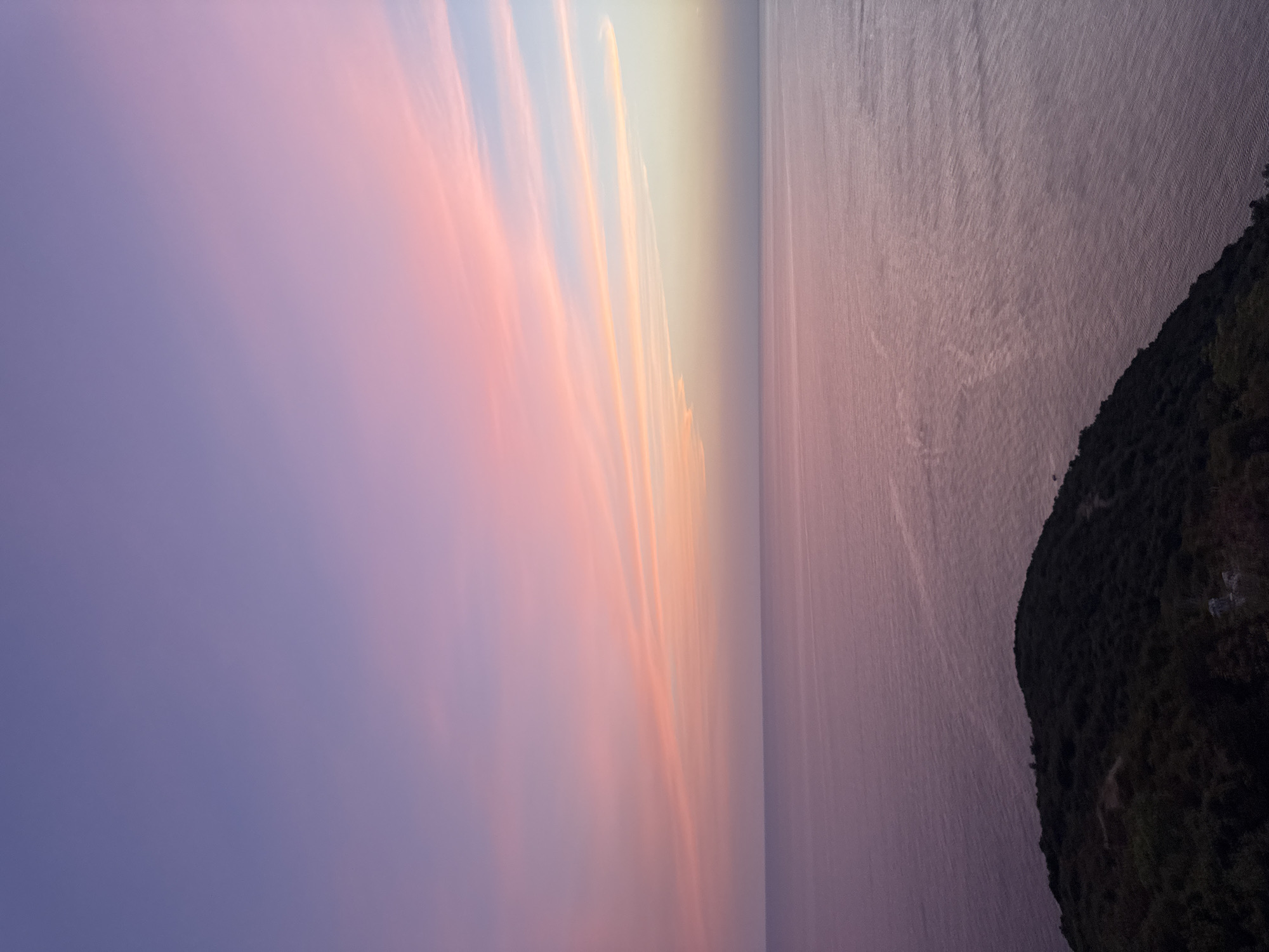 A dusky sky transitions from a deep lavender at the top to soft pinks and oranges near the horizon. Wispy clouds streak across the upper portion of the image, catching the fading light. Below the sky, a vast expanse of calm ocean stretches out, its surface reflecting the muted colors of the sky in subtle shades of purple and pink. In the foreground, a dark, tree-covered hillside juts out into the water from the left side of the frame, its vegetation creating a stark contrast against the soft hues of the sea and sky. The scene suggests a tranquil coastal landscape at either sunrise or sunset. The location is not within the city of Zagora, Montenegro; it is a general coastal landscape.