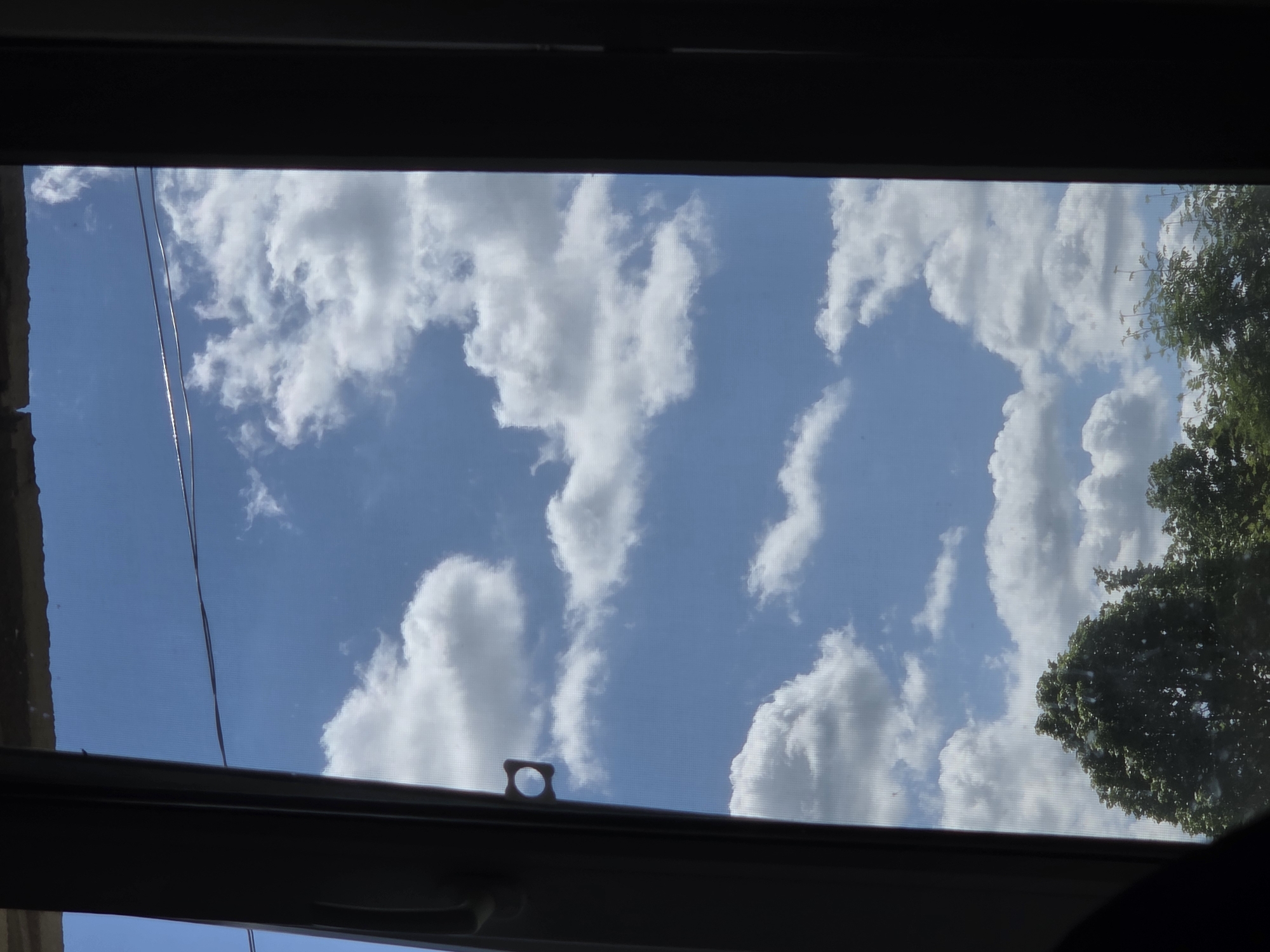 The image depicts a view of the sky through a window. The sky is blue with scattered white clouds. Part of a tree is visible on the right side of the window. The window frame is dark and rectangular. Two wires or strings run parallel to each other across the left side of the window. An object with a circular hole is attached to the lower edge of the window frame. The location is Unknown, Unknown.